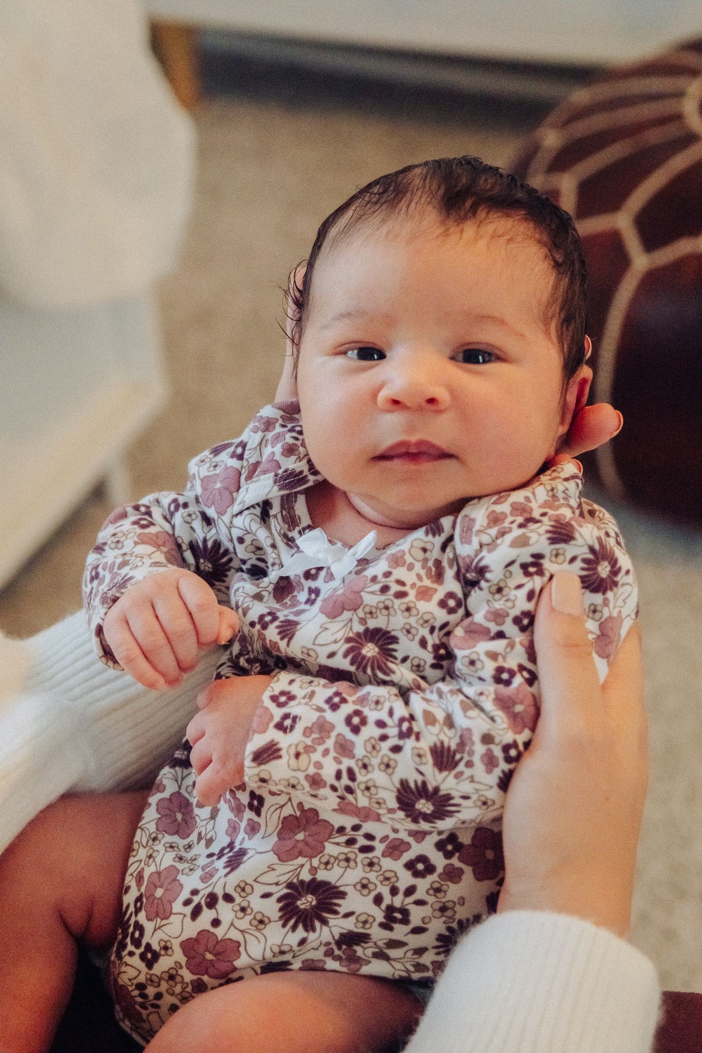 Newborn baby in floral romper being held by adult hands in a cozy indoor setting.