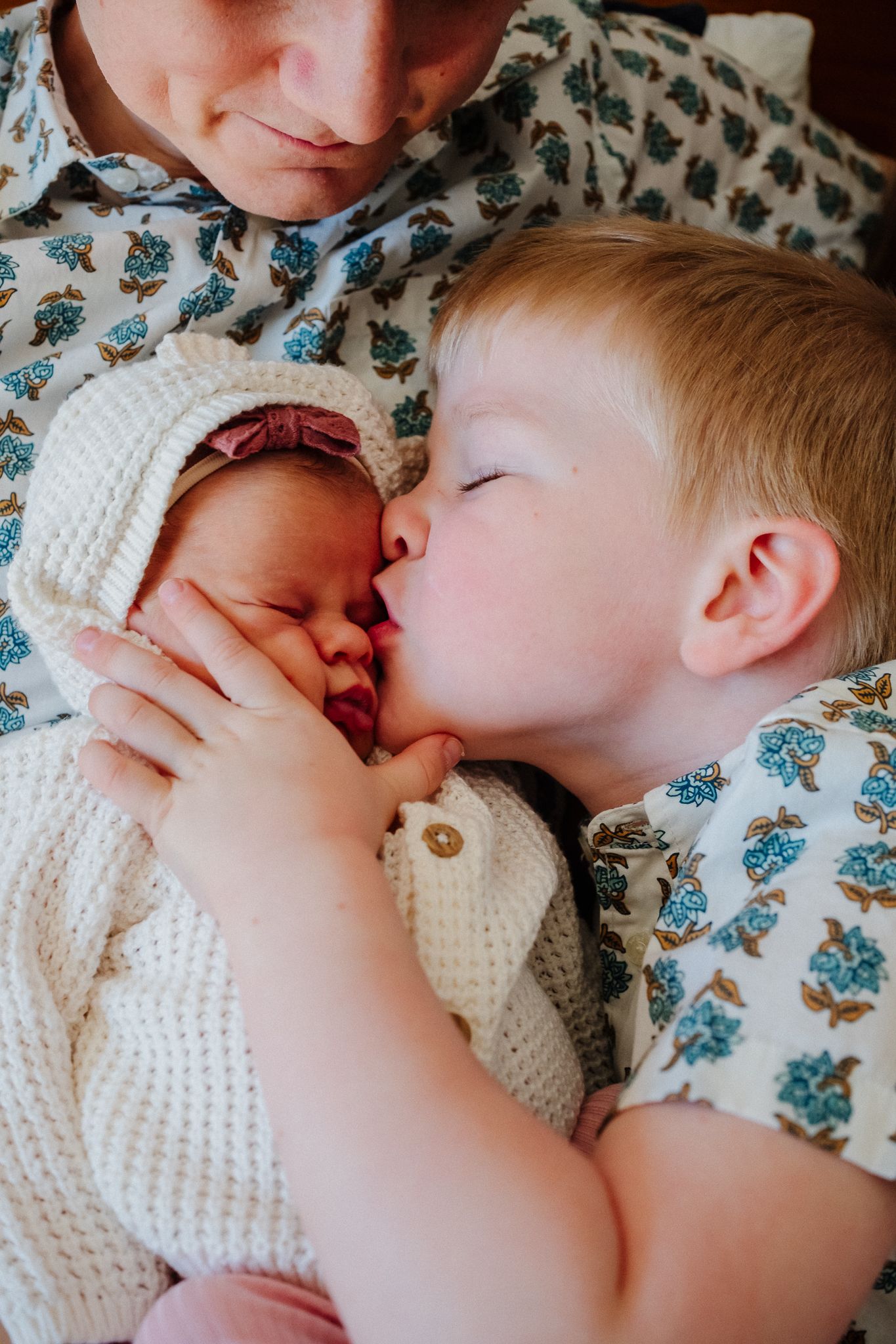 Toddler boy gently kissing newborn baby sibling while mother holds infant in cozy knit outfit.
