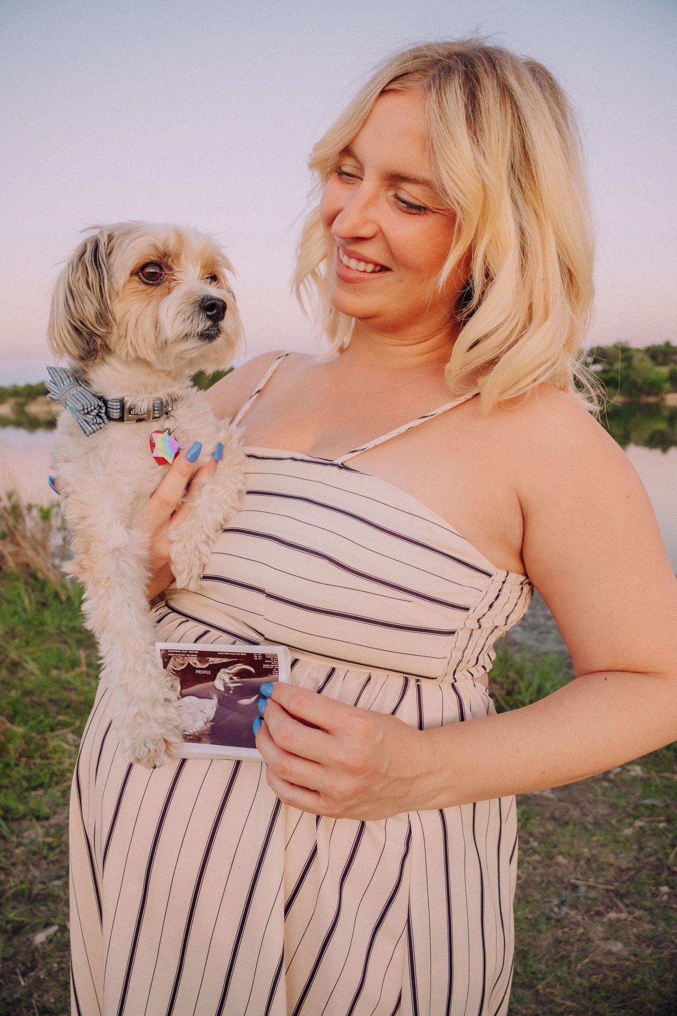 A person in a striped sundress holds a small scruffy white dog outdoors during golden hour.