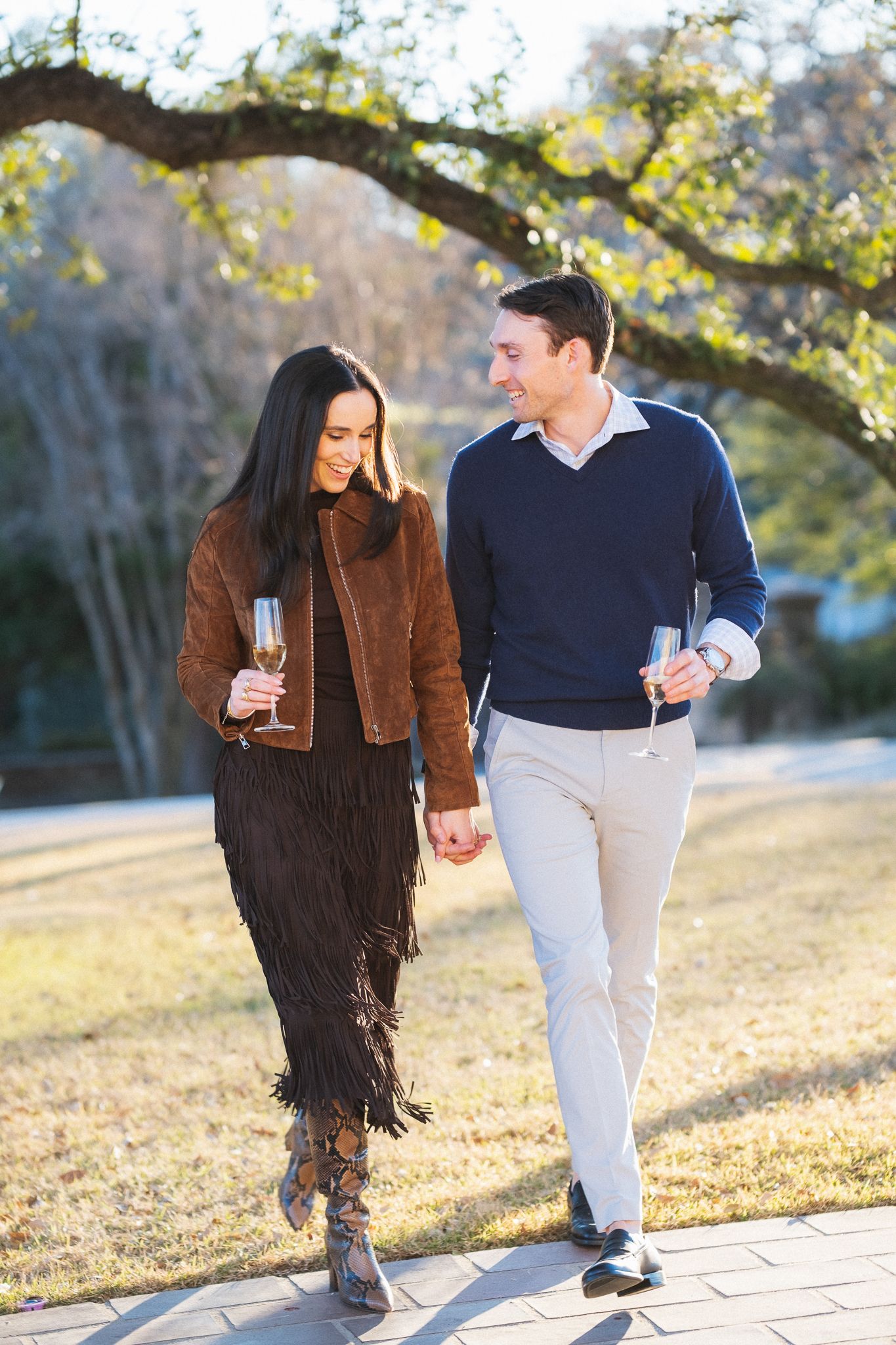 Couple walking outdoors holding champagne glasses, woman in brown fringe jacket, man in navy sweater and khakis.