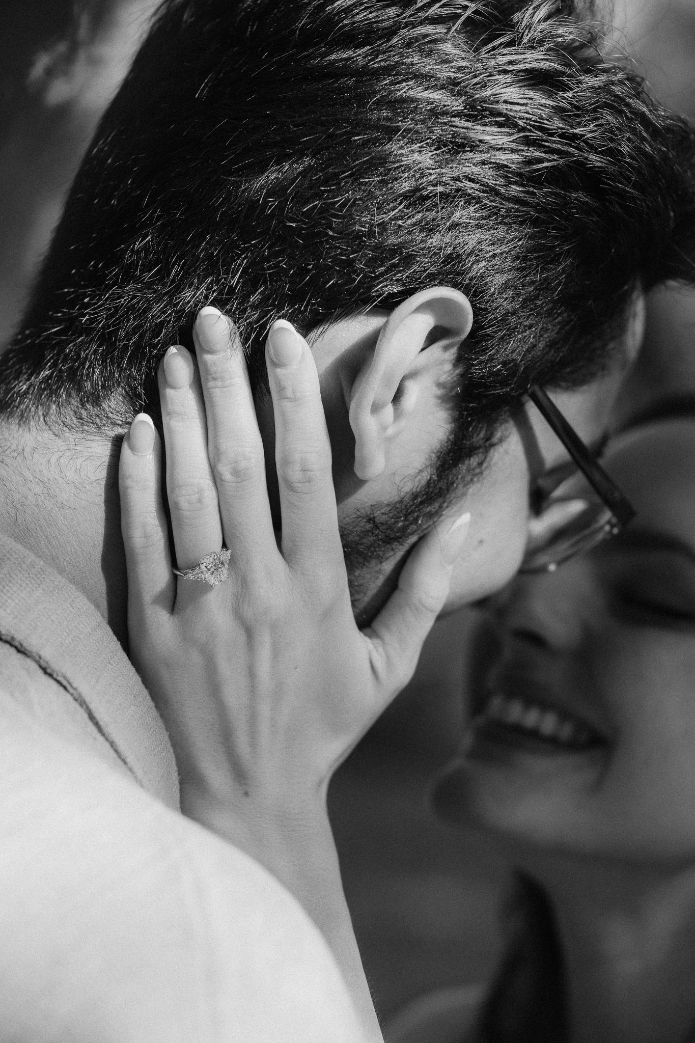 Black and white photo of a couple embracing, woman with ring gently holding mans face, smiling softly.