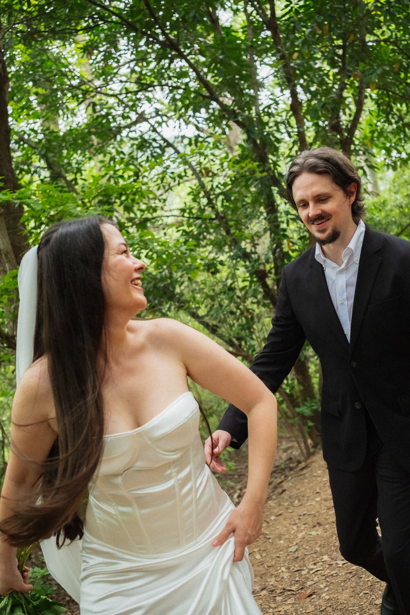 A smiling bride in a white strapless gown holds hands with a groom in a black suit among lush green trees.