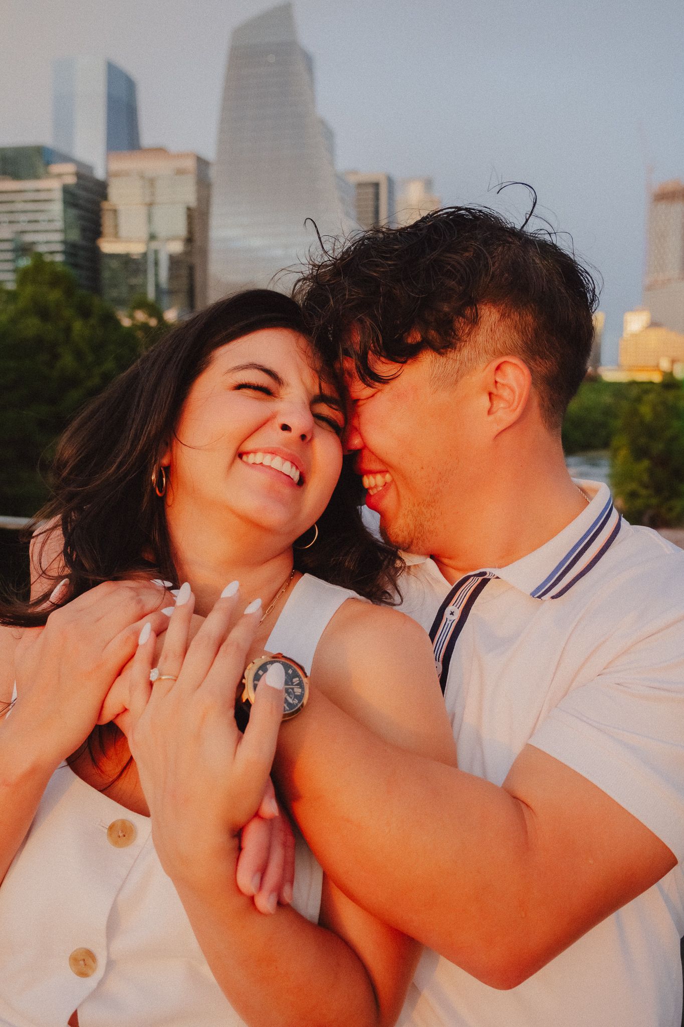 Happy couple embracing at sunset with city skyline backdrop, woman showing engagement ring while man kisses her cheek.