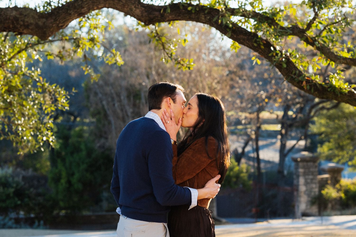 Couple sharing a romantic kiss outdoors under a large oak tree with golden sunlight filtering through the leaves.