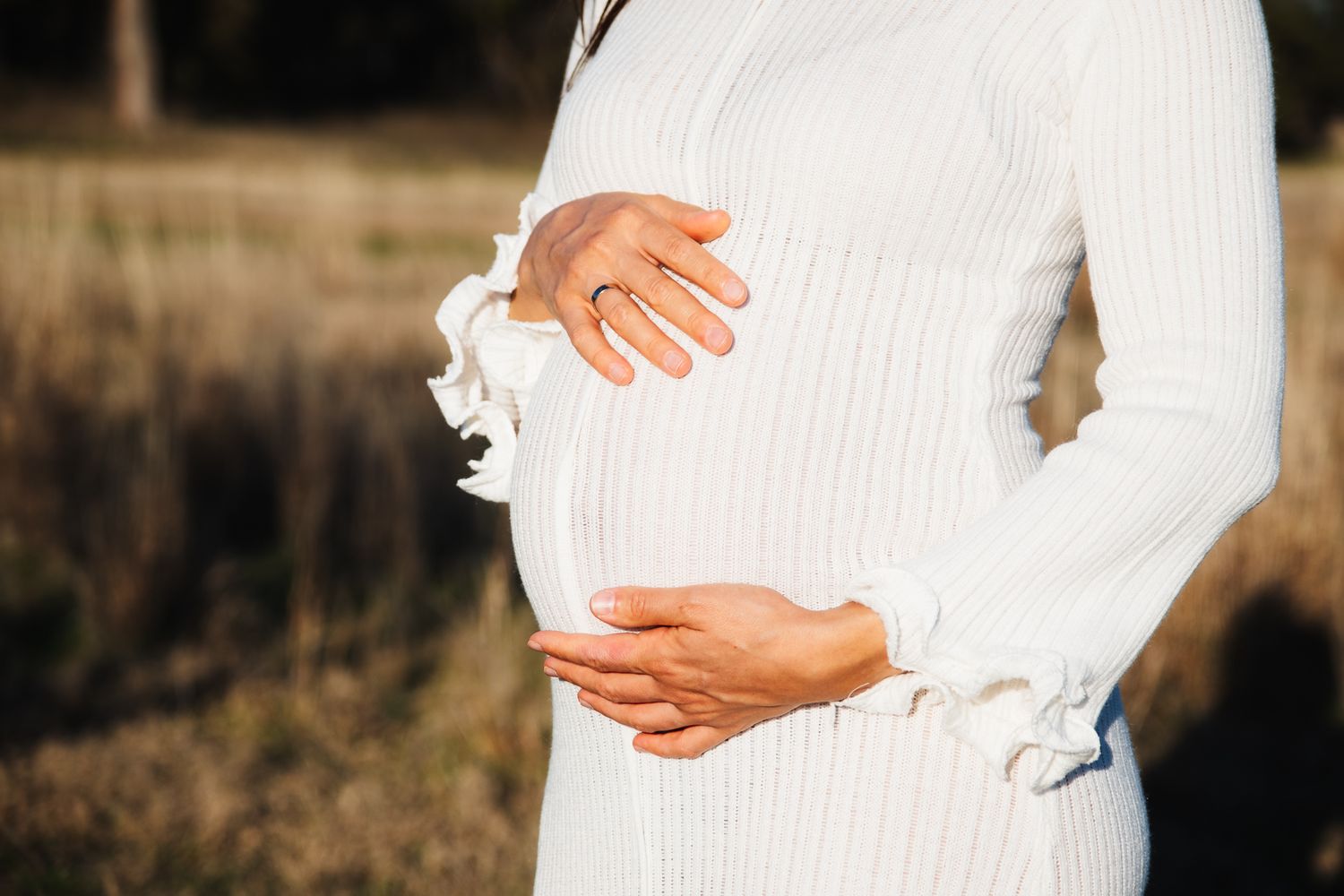 Person wearing a white ribbed sweater cradles their pregnant belly in sunlit outdoor setting.