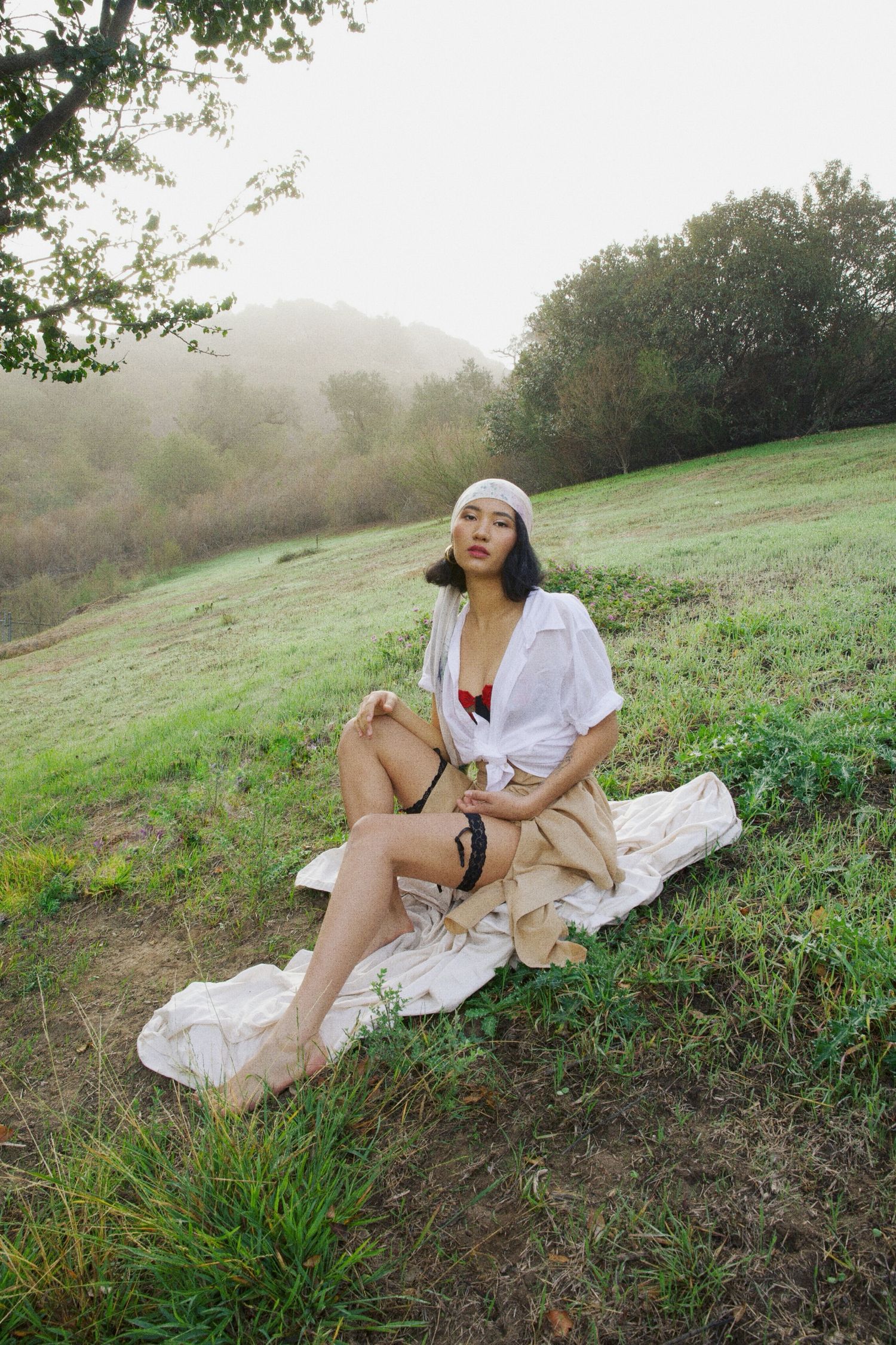 Person in white shirt and beige shorts sitting on picnic blanket on grassy hillside during misty morning.