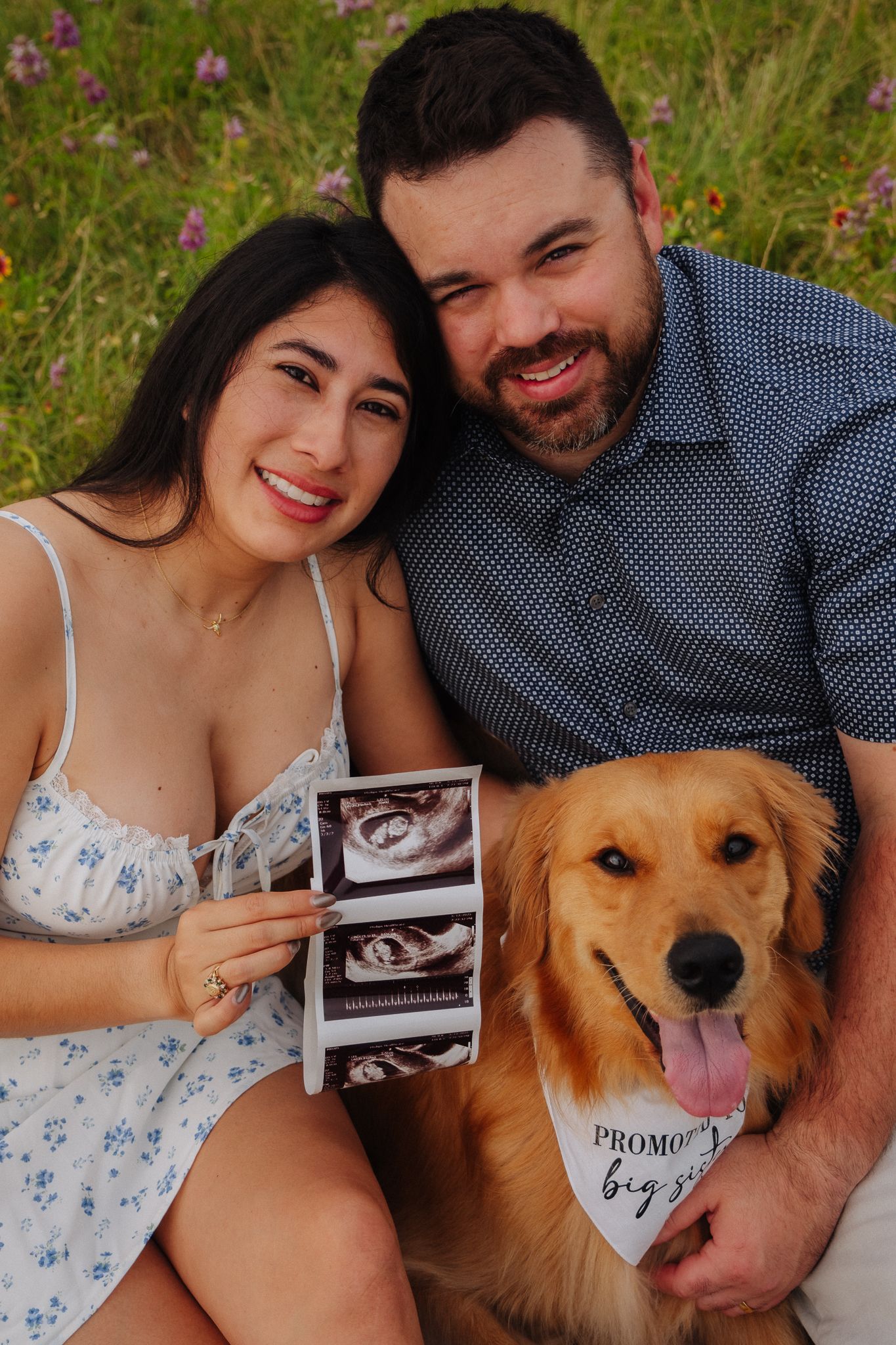 A couple sits outdoors with their golden retriever dog while displaying ultrasound photos in a pregnancy announcement.