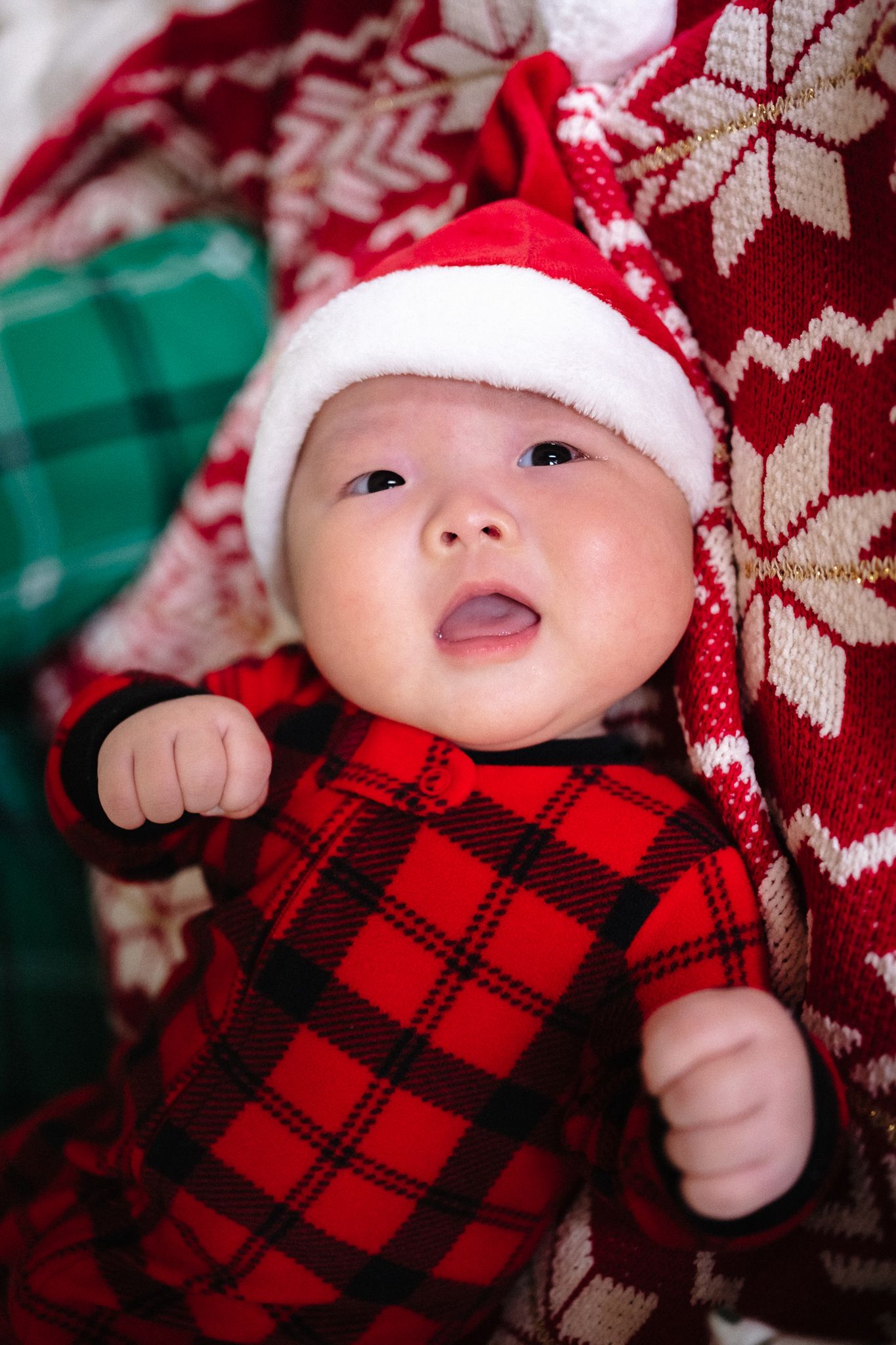 Adorable baby in red plaid onesie and Santa hat surrounded by festive Christmas blankets.