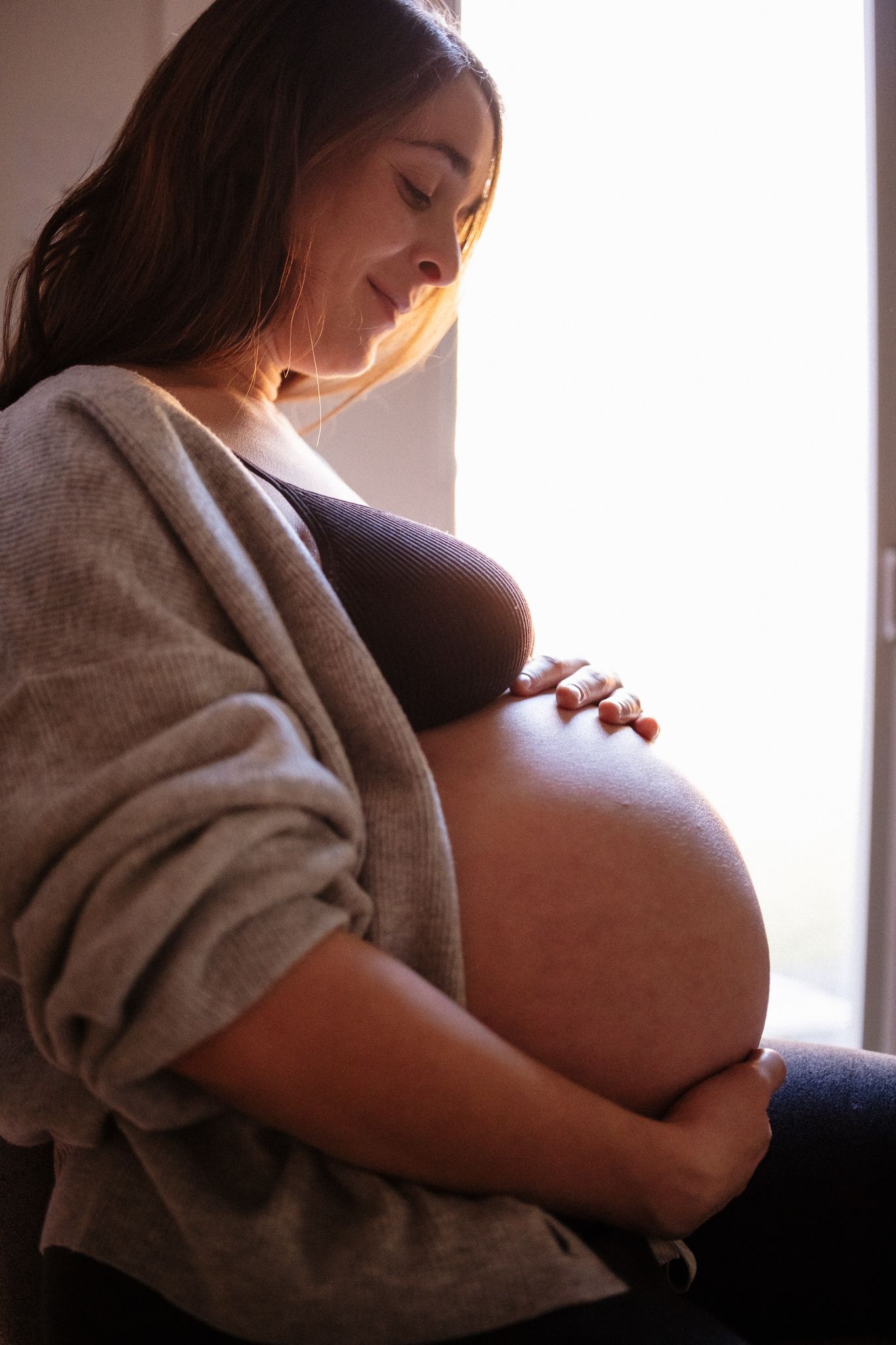 An expectant mother in a grey sweater gazes down at her pregnant belly in warm window light.