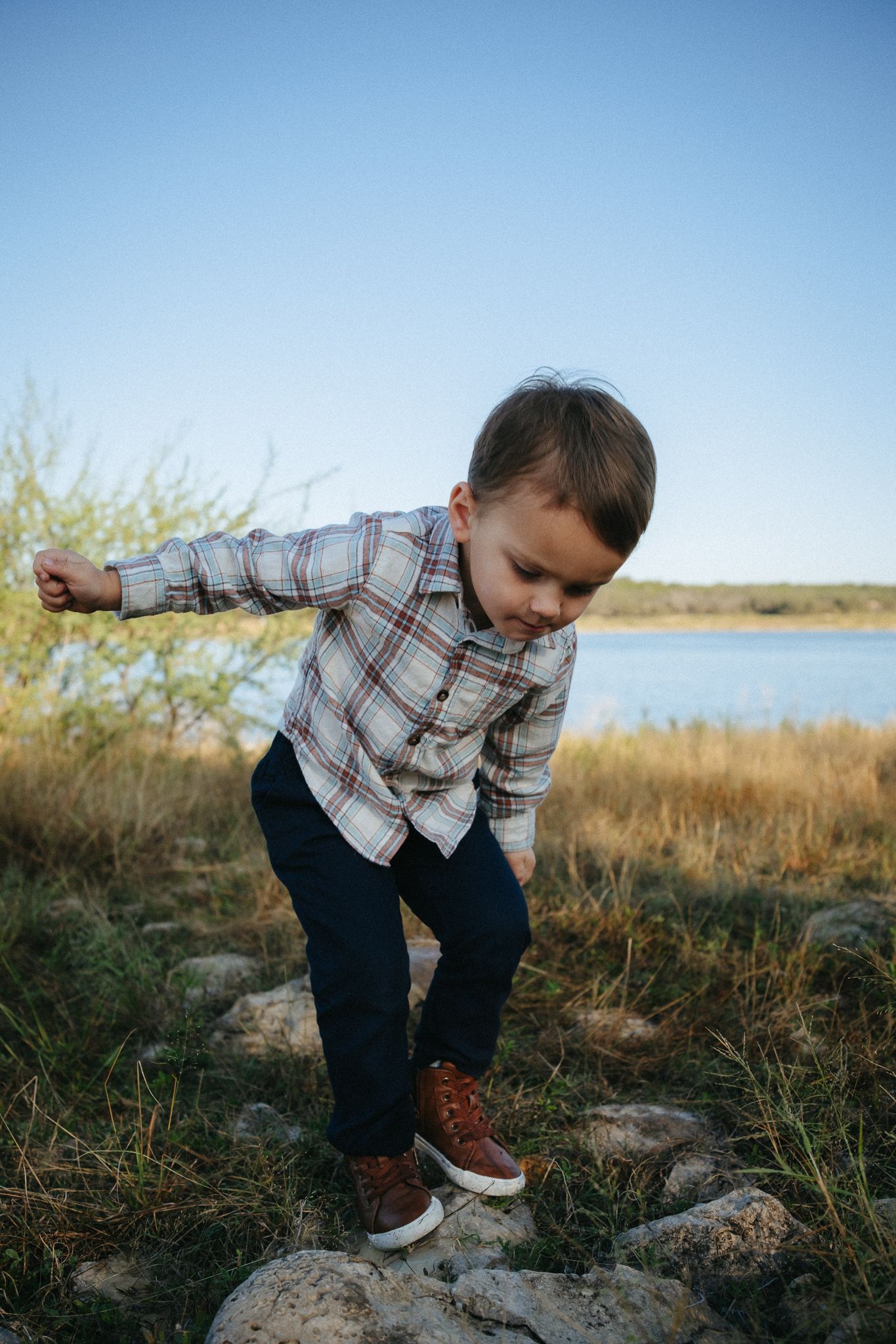 Small child in plaid shirt and dark pants balances on rocks near a lake during golden hour.