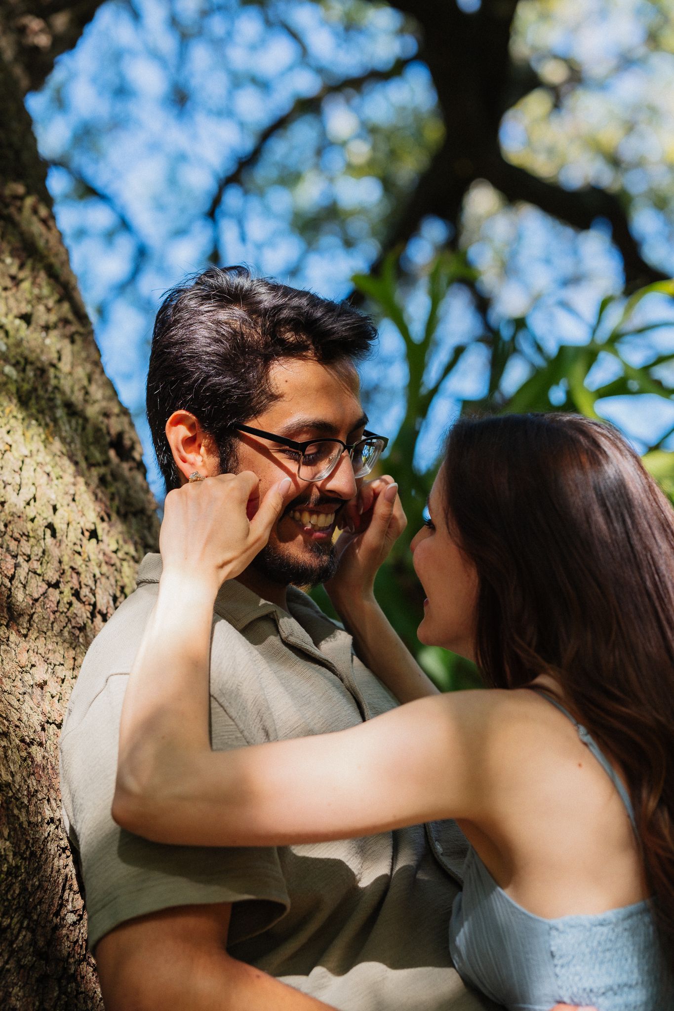 A couple shares a tender moment under a tree, woman gently cupping man's face as they smile at each other.