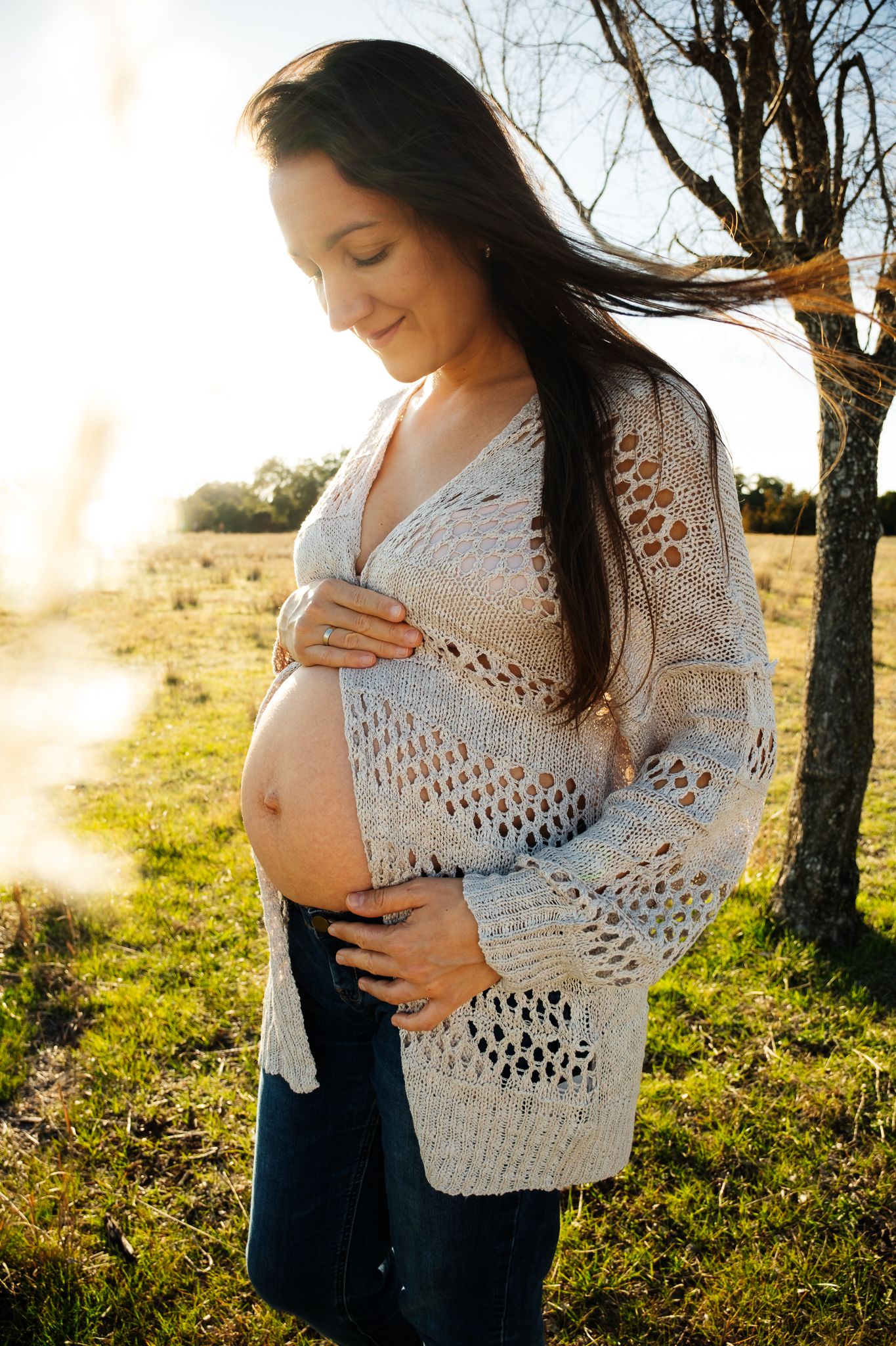 A pregnant woman in a white crochet sweater stands in a sunlit field at golden hour with bare trees in background.