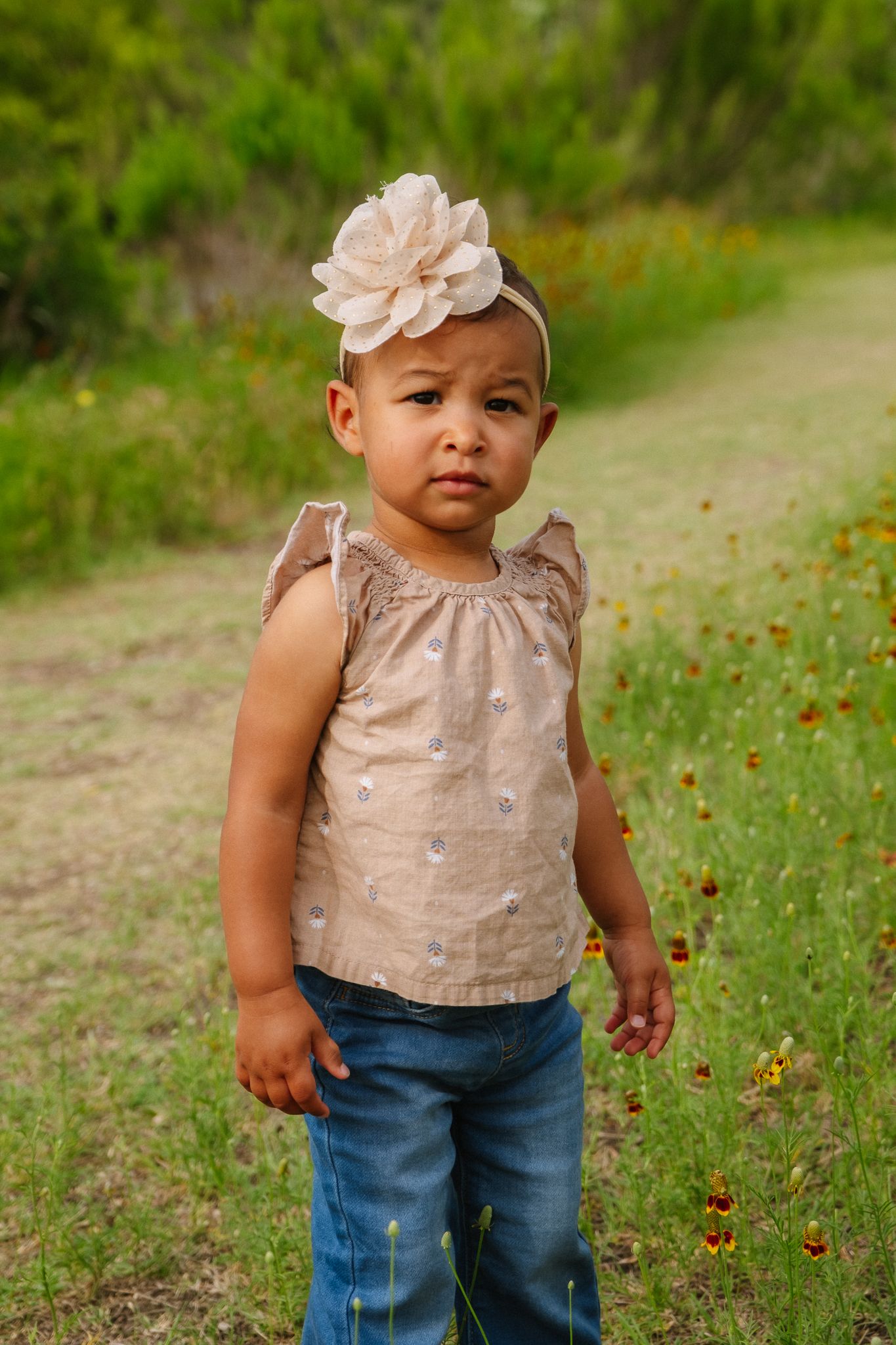 A toddler wearing a beige sleeveless top and blue jeans stands in a grassy outdoor setting with a floral hair accessory.