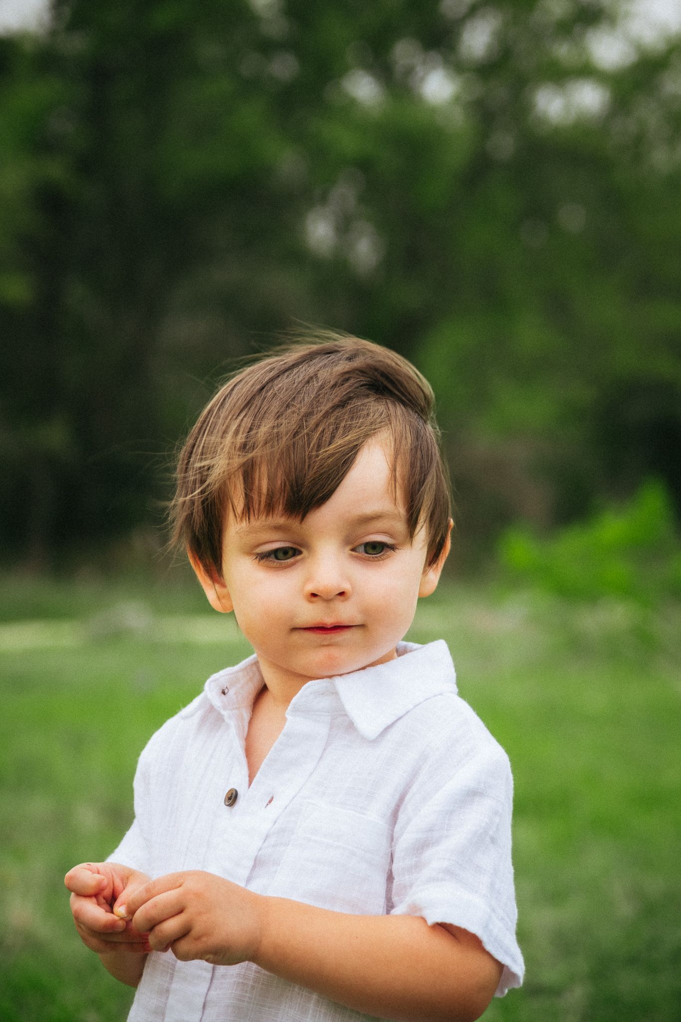 Young child in white shirt poses naturally outdoors against green woodland background in summer.