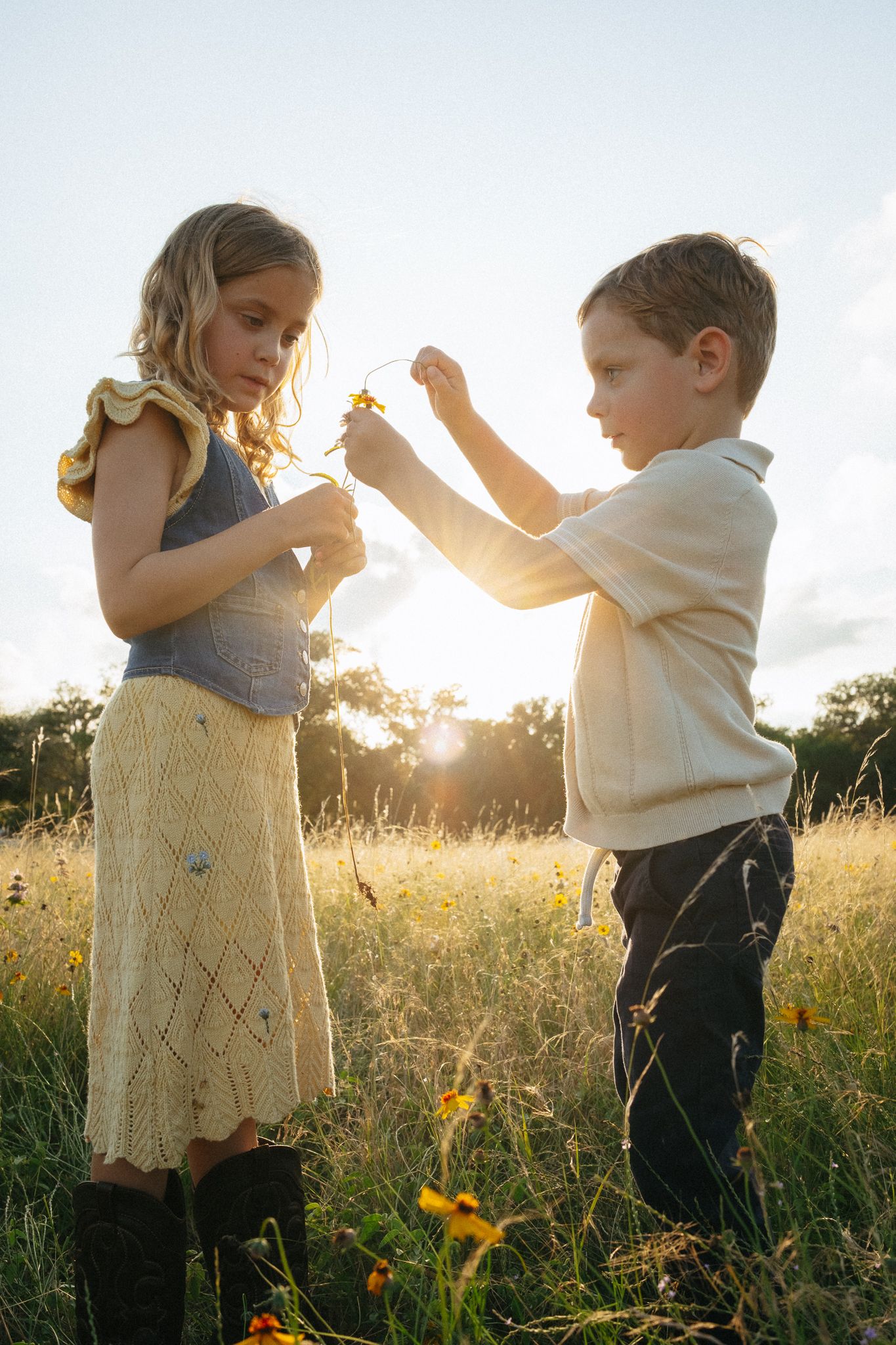 Two children examine wildflowers in a sunlit meadow at golden hour, girl in yellow skirt and boy in cream shirt.