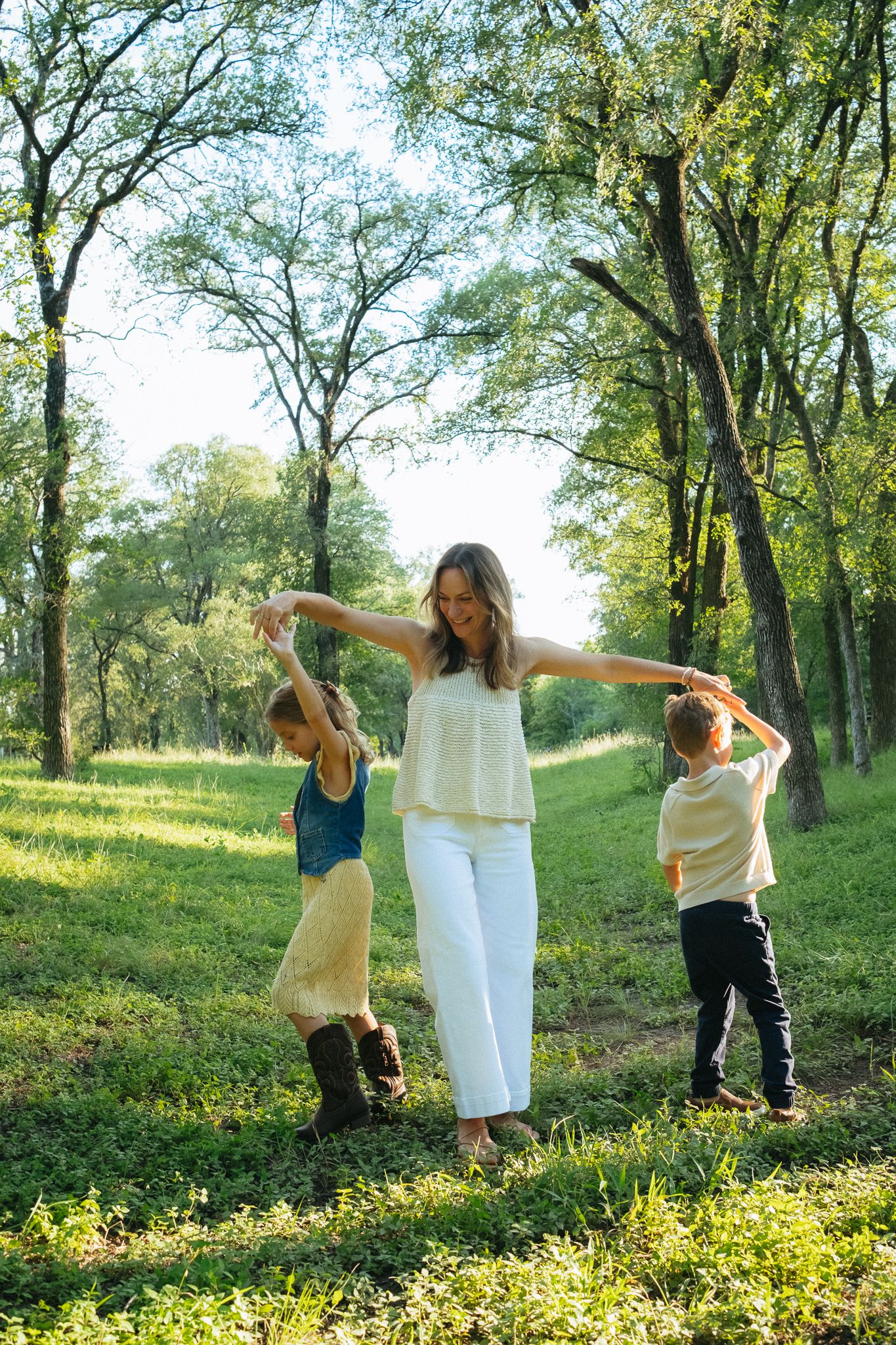 A woman in white holds hands with two children while dancing outdoors in a sunlit green forest clearing.