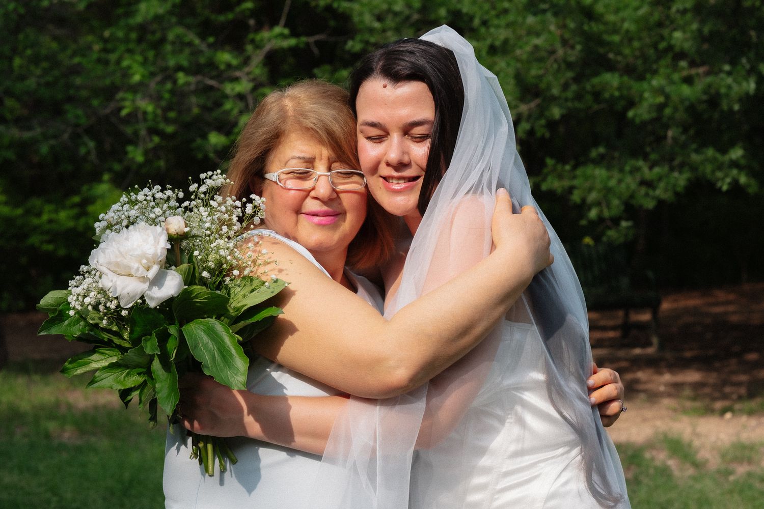 Bride in white gown and veil hugging older woman holding white bouquet outdoors at wedding celebration.