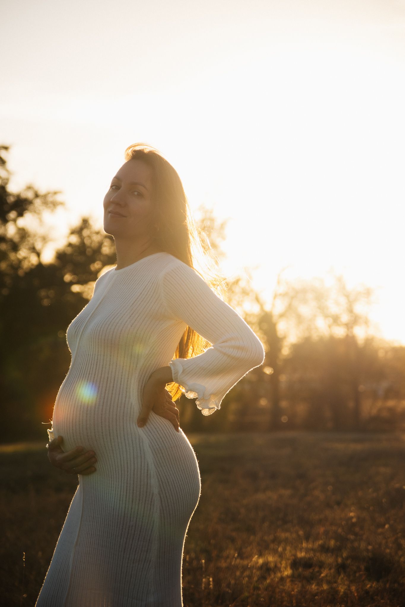 A pregnant person in a white dress poses in golden sunlight during sunset in a field, creating a dreamy silhouette.
