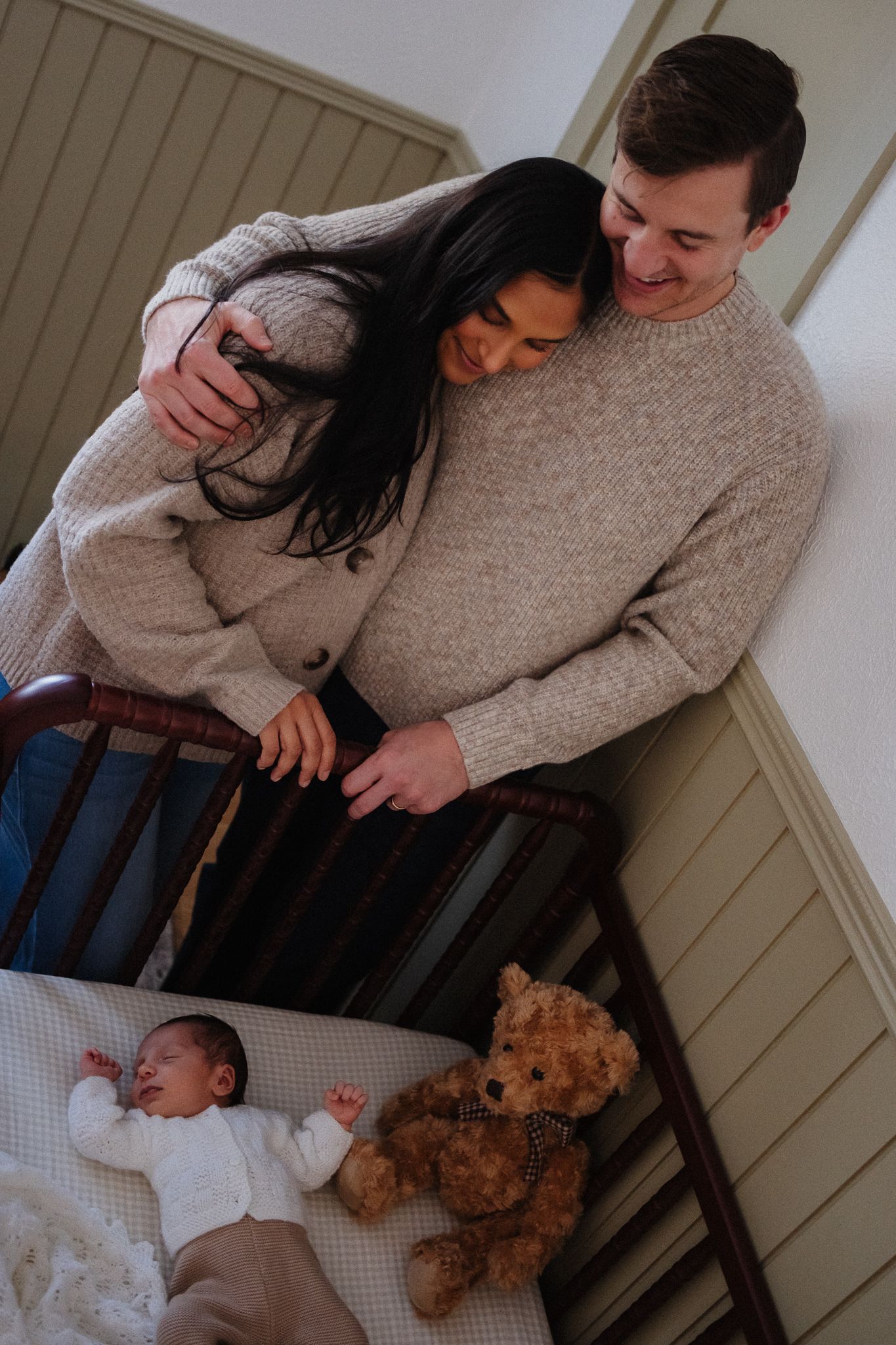 Parents in neutral sweaters cuddle their baby while sitting together with a teddy bear nearby.