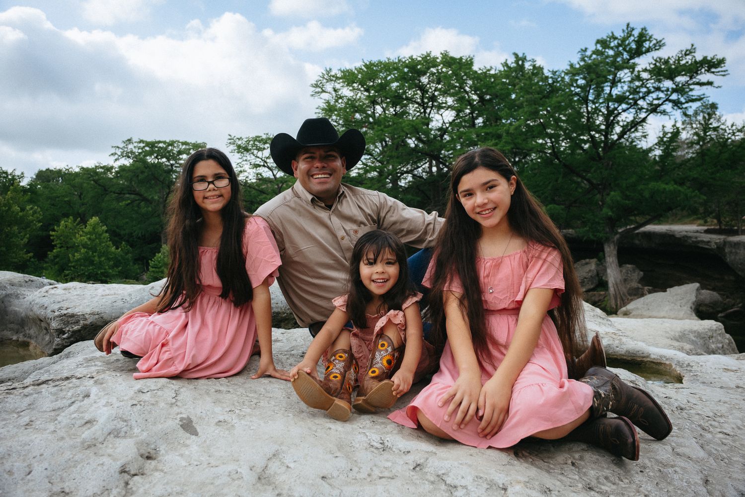 Family of five posing on rocks outdoors, with three girls in pink dresses and a man in a cowboy hat, surrounded by trees.