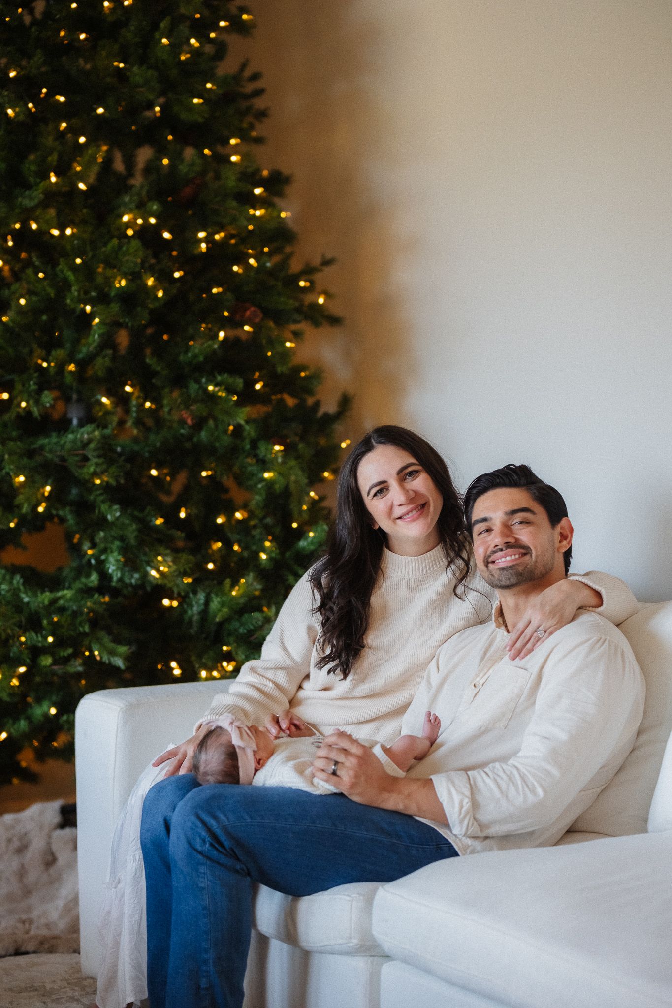 Couple in matching cream sweaters sitting on white sofa embracing in front of lit Christmas tree.