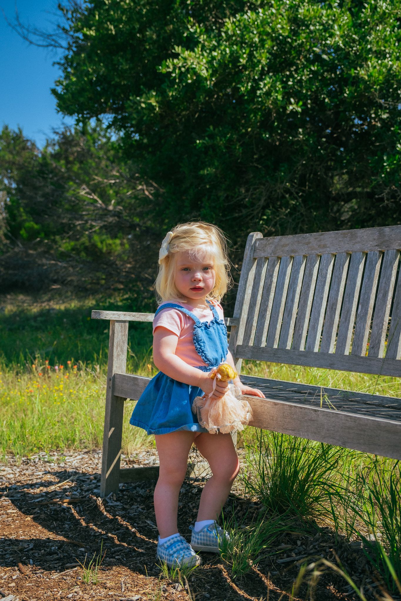 A small child sits and plays on a wooden bench surrounded by lush greenery in a park setting.