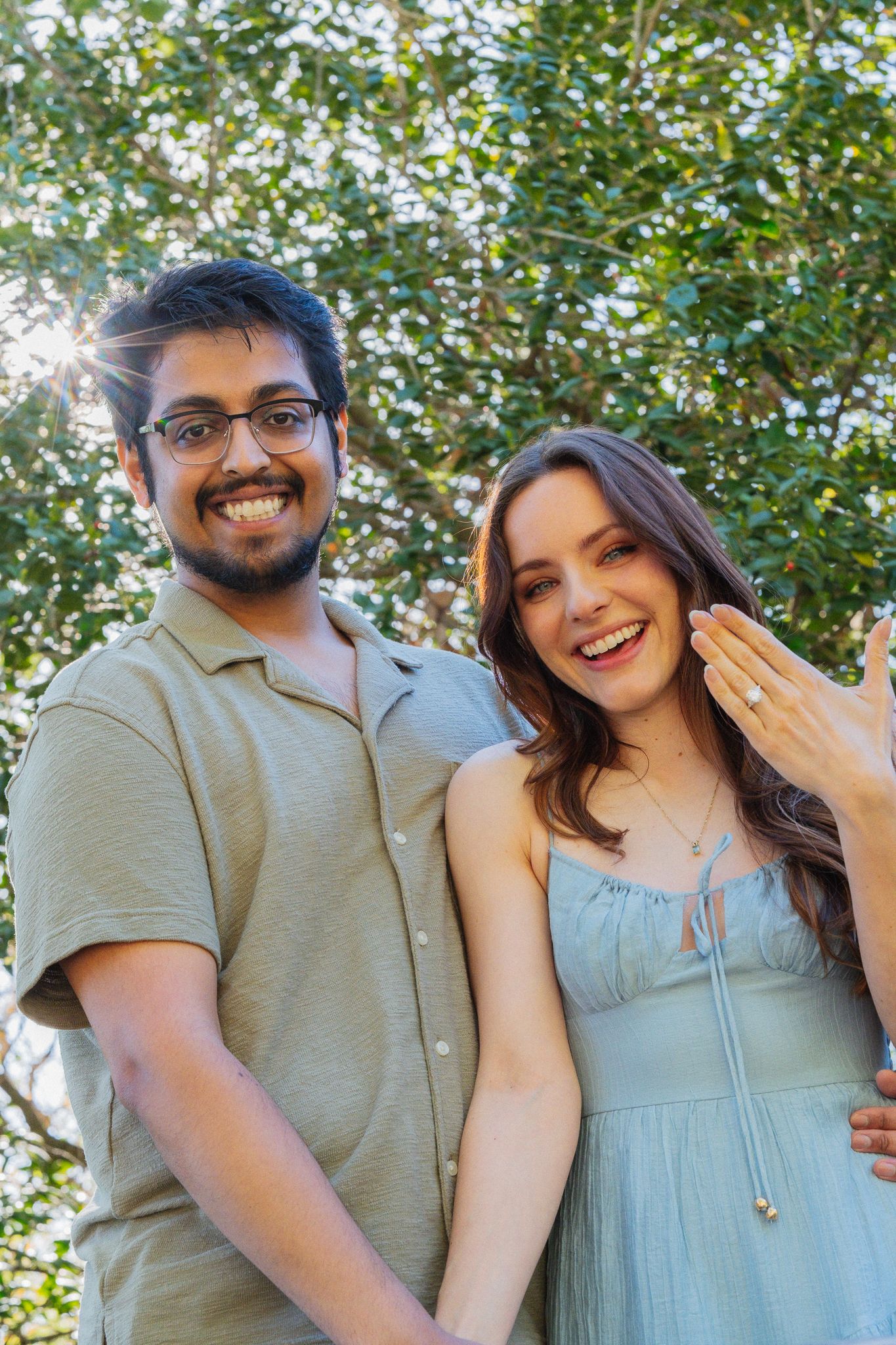Happy couple smiling outdoors, woman showing engagement ring, man in glasses, green trees background.