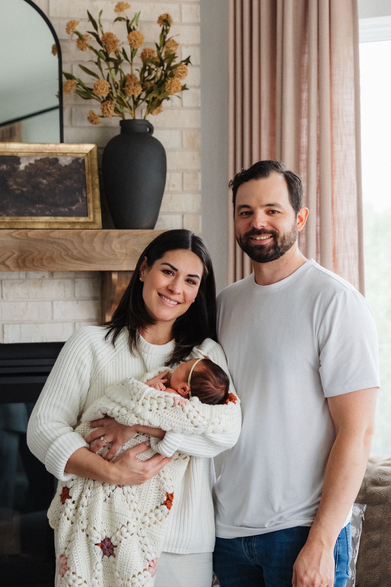 Smiling couple holding their newborn baby in a cozy living room with fireplace and floral decor.