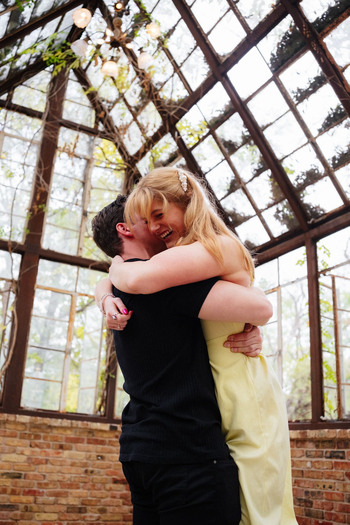 Couple embracing and laughing in a rustic greenhouse with brick walls and glass ceiling.