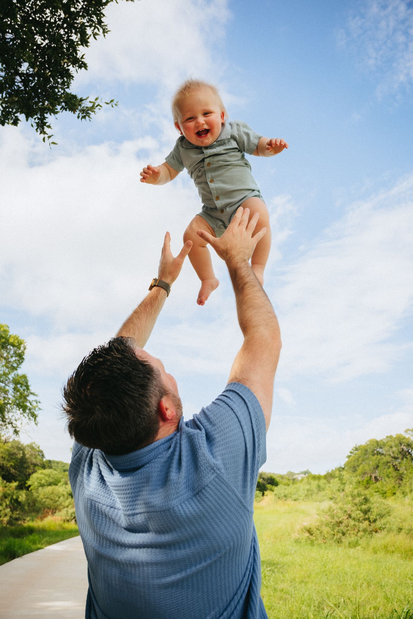 A person in a blue shirt lifts a small child wearing grey clothing high into the air against a bright blue sky.
