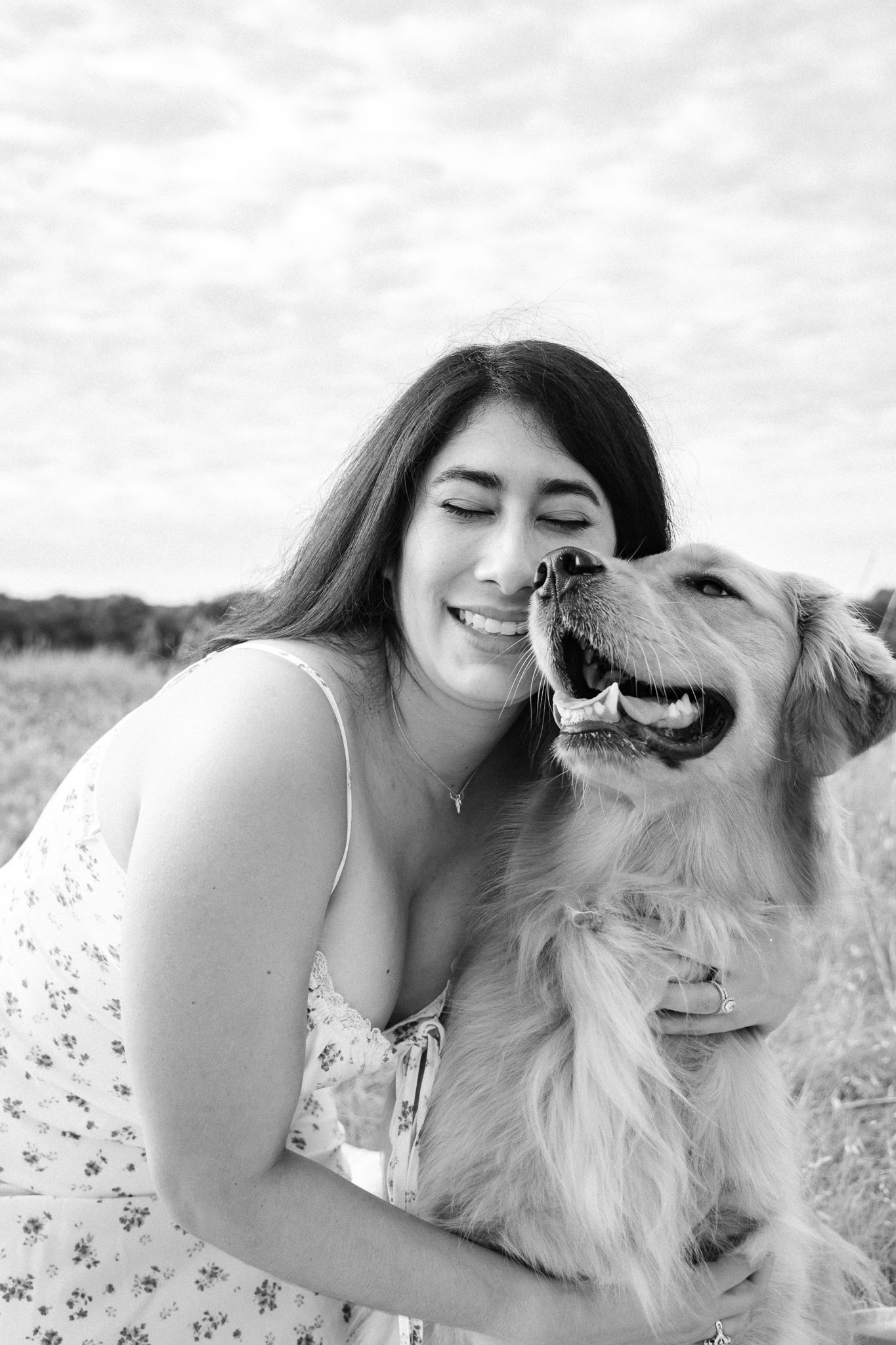 Black and white photo showing a happy golden retriever and person in a floral dress sharing a joyful outdoor moment together.