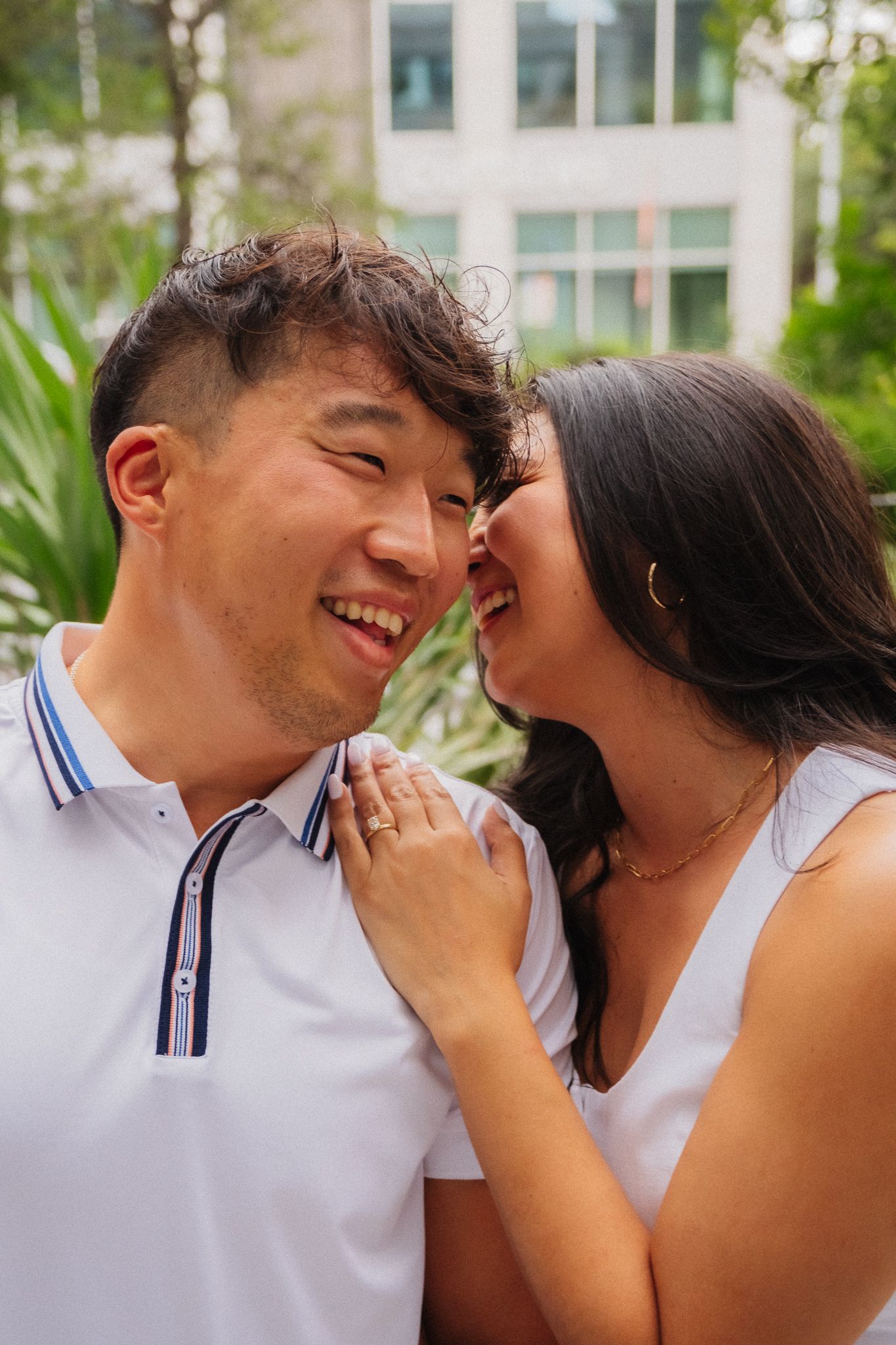 A joyful Asian couple in white outfits share a tender moment outdoors, with the woman whispering to the laughing man.