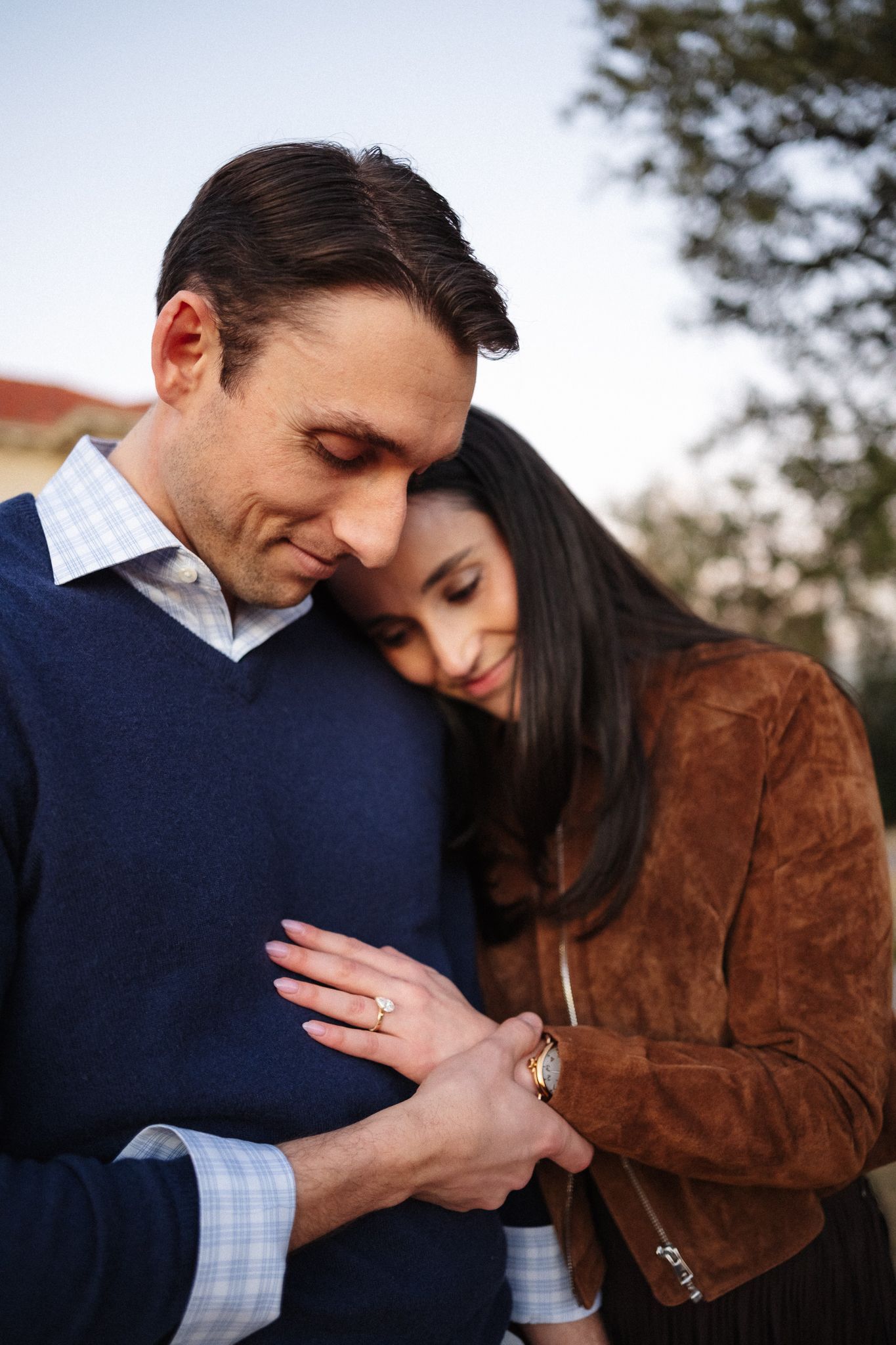 A couple shares a tender embrace outdoors, woman in brown jacket resting head on mans shoulder, wearing a gold ring.