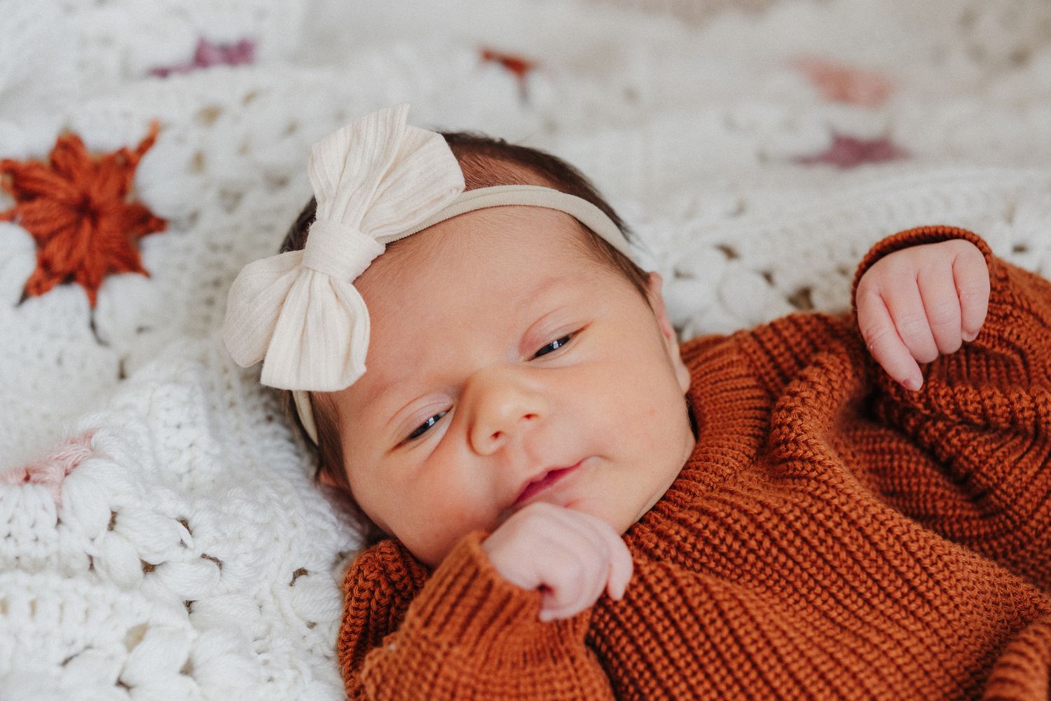 Newborn baby girl in rust orange knit outfit and white bow headband lying on white crochet blanket.