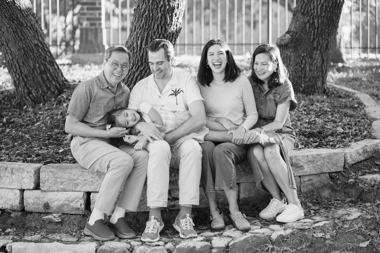 Family sitting together on grass beneath trees in a casual black and white outdoor portrait session.