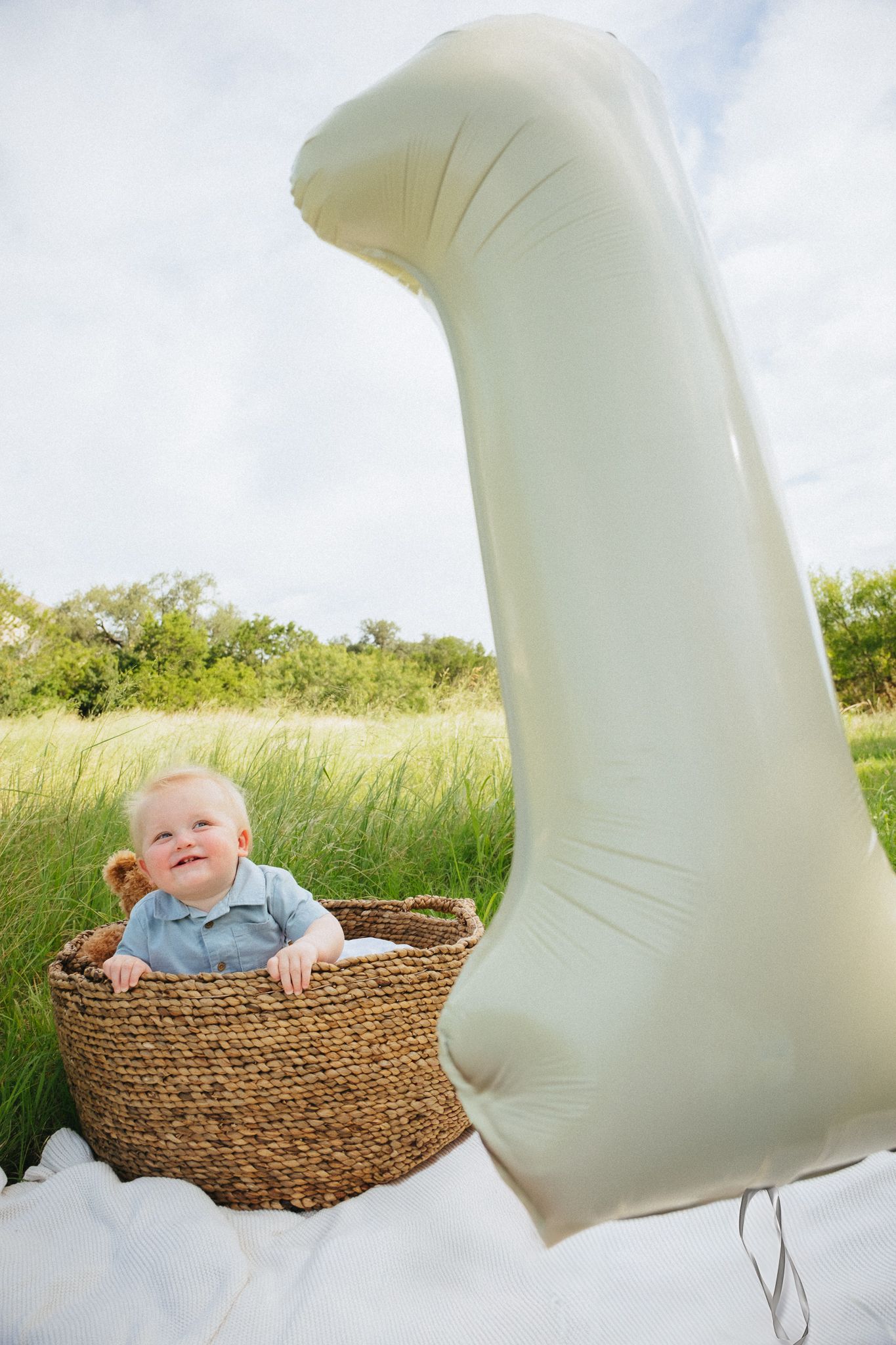 A baby lies in a woven basket outdoors with large white angel wings prop against a natural background.