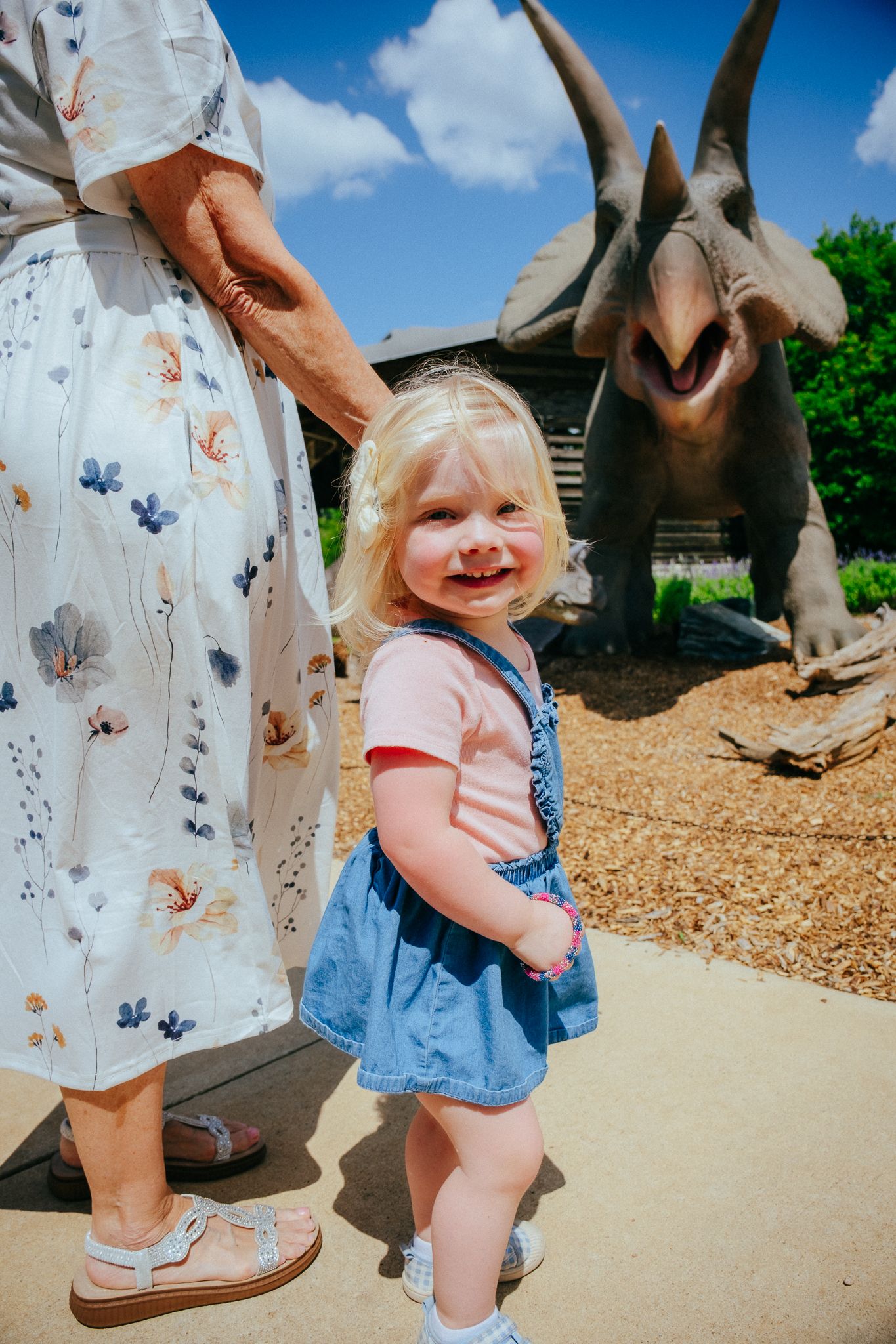 Small child in denim overalls visiting a dinosaur exhibit at an outdoor park on a sunny day.