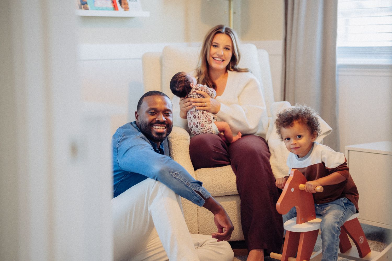 Happy family of four posing in nursery, parents smiling with newborn baby and toddler on rocking horse.