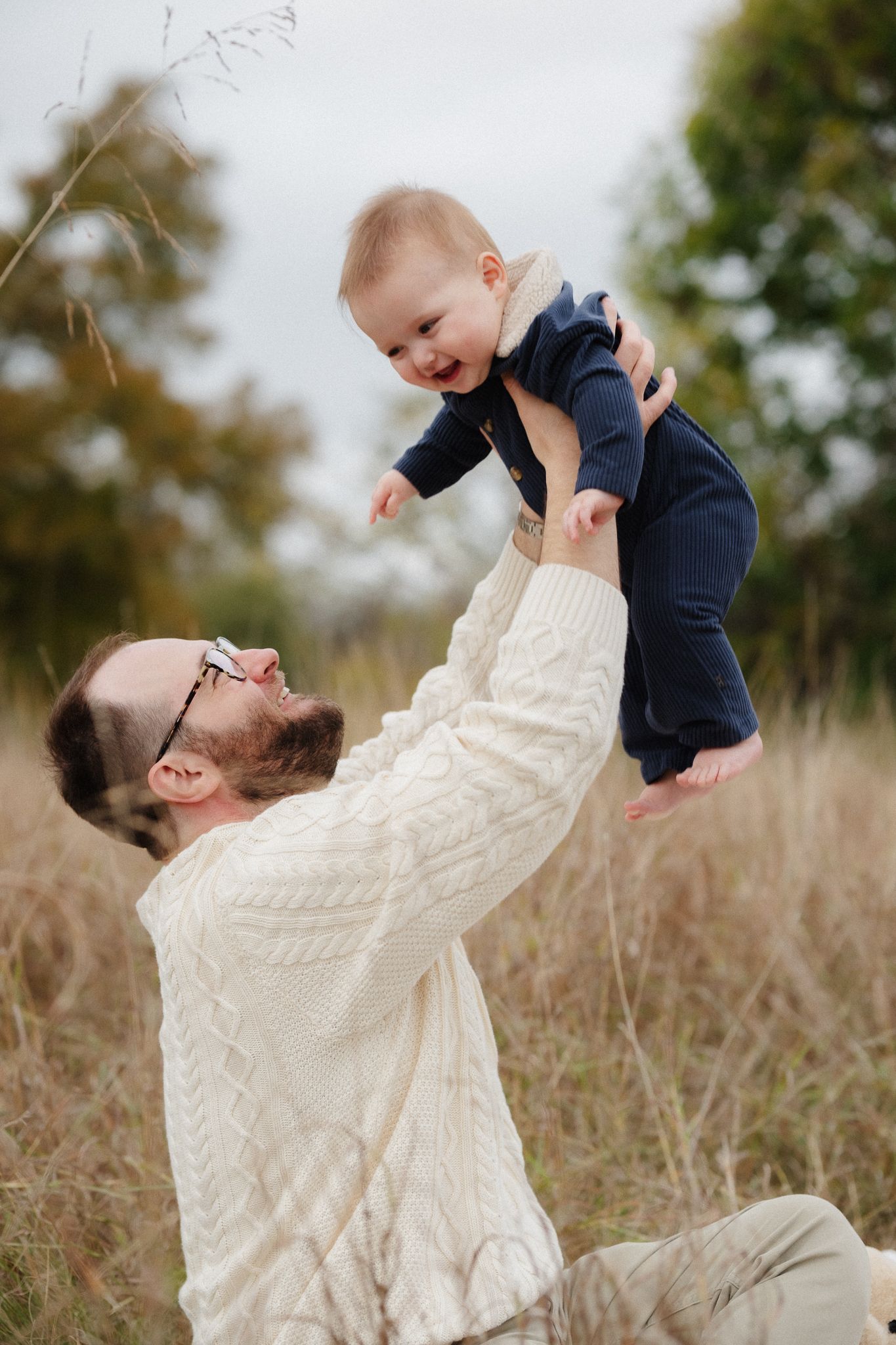 A parent lying in tall grass lifts and plays with a small child wearing navy blue clothing against a natural outdoor backdrop.