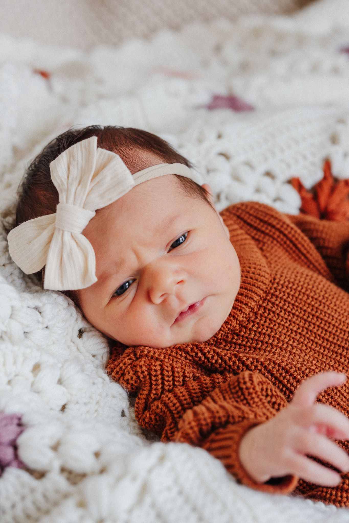 Newborn baby girl in rust knit romper and white bow headband lying on white crochet blanket.