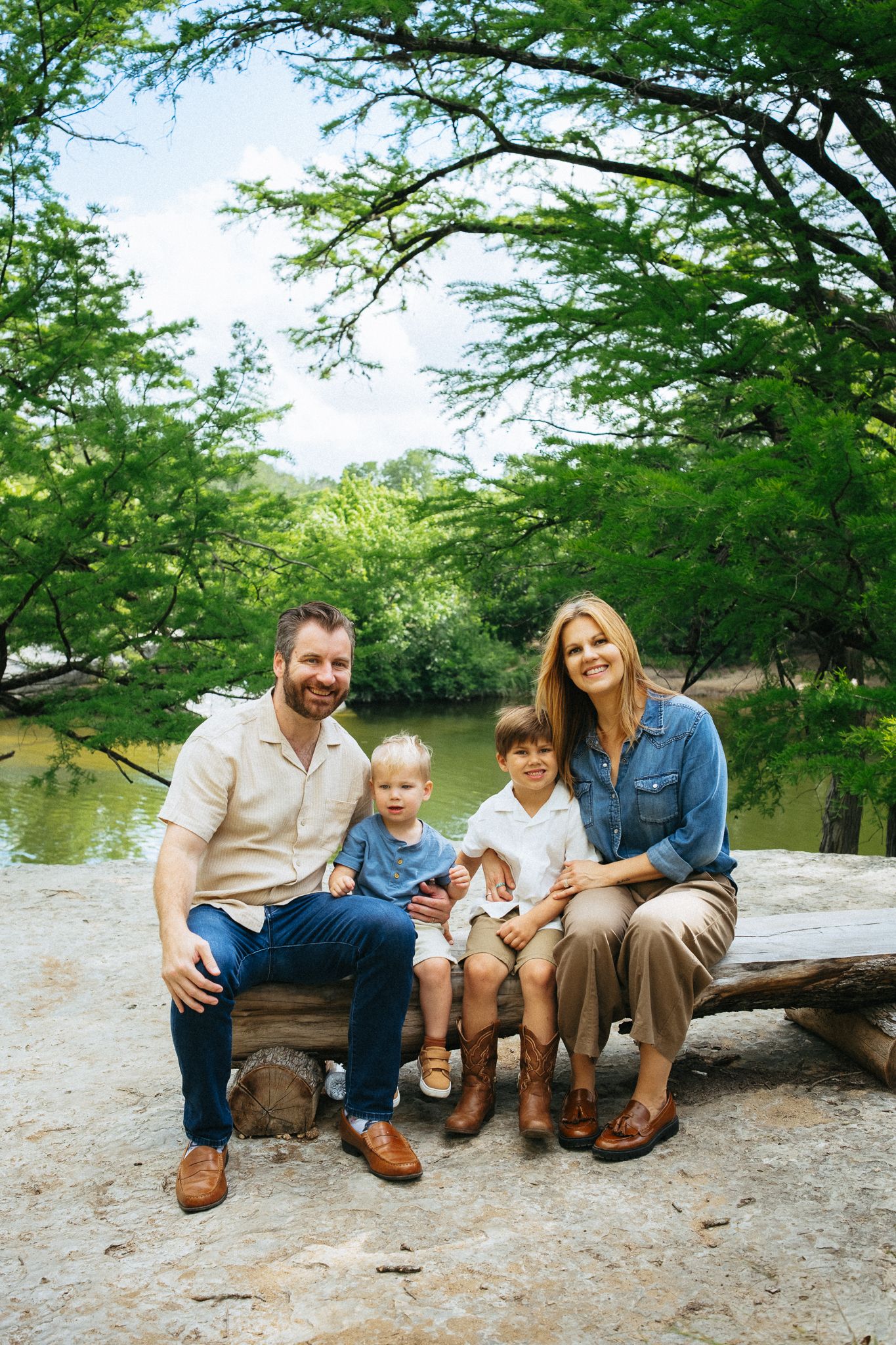 Family of four smiling outdoors near a green tree-lined river, seated on a log in casual attire.