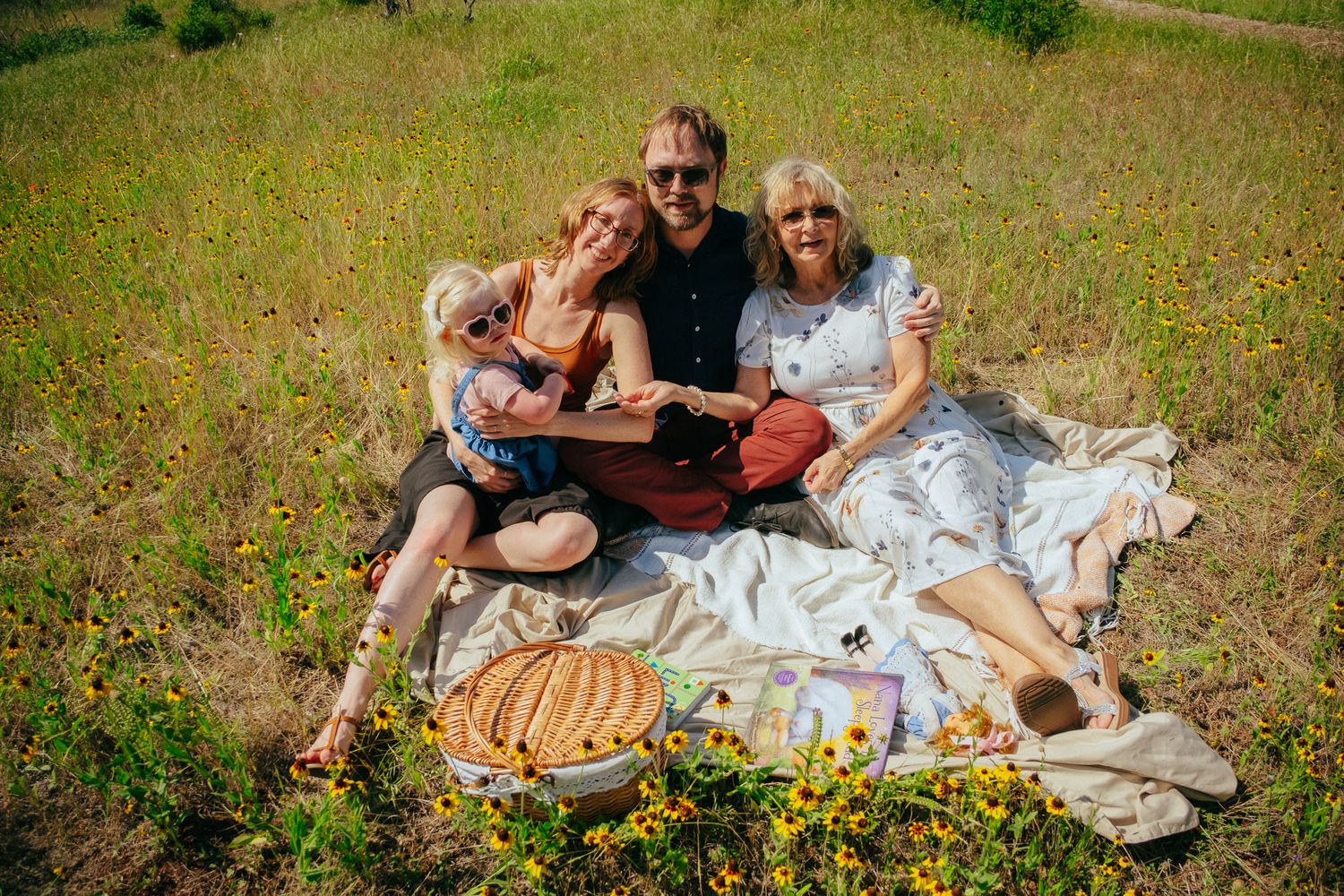 Family of four enjoying a sunny outdoor picnic on a blanket in a wildflower field with food and drinks.
