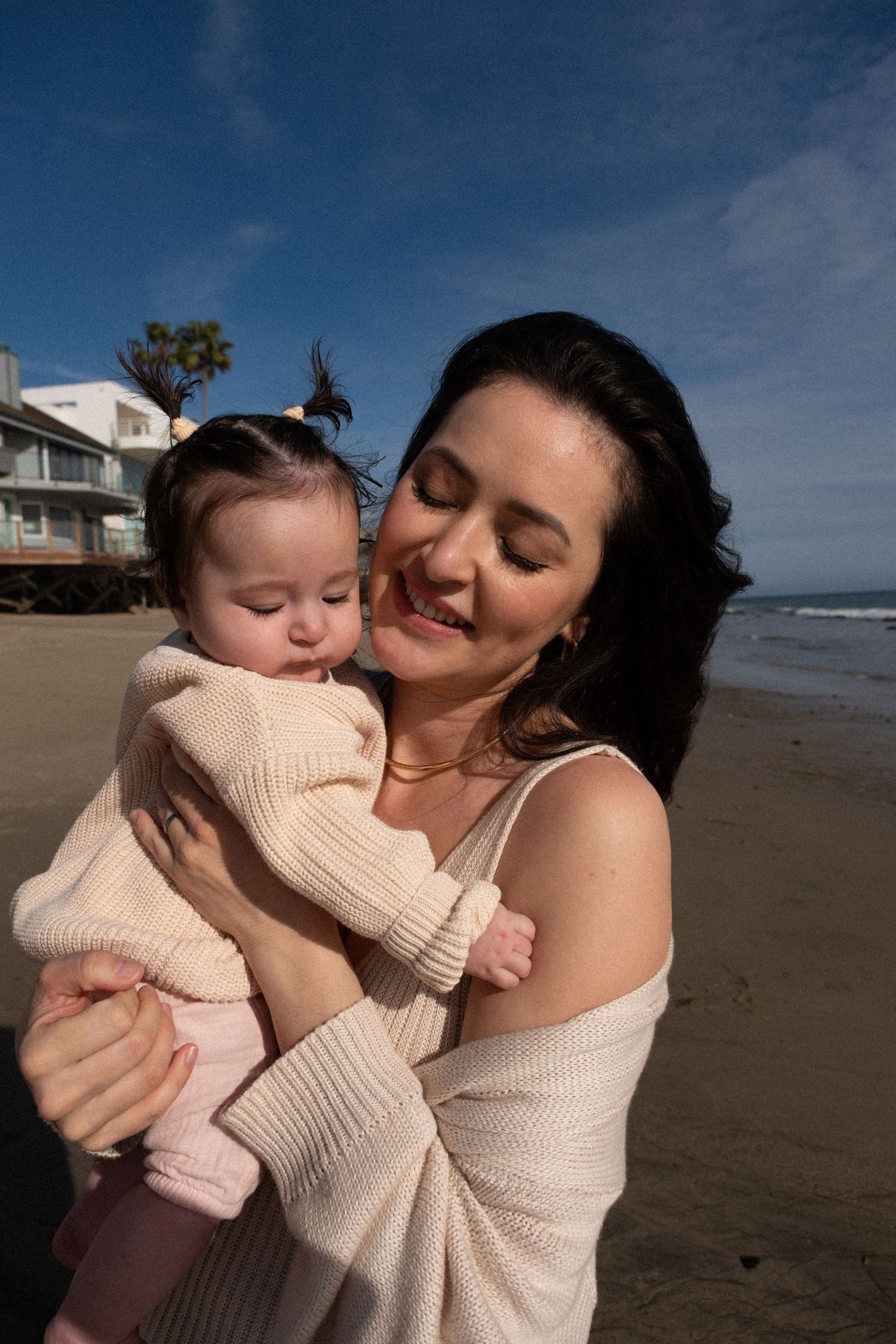 A tender moment between parent and baby captured on a sunny beach with coastal homes in the background.