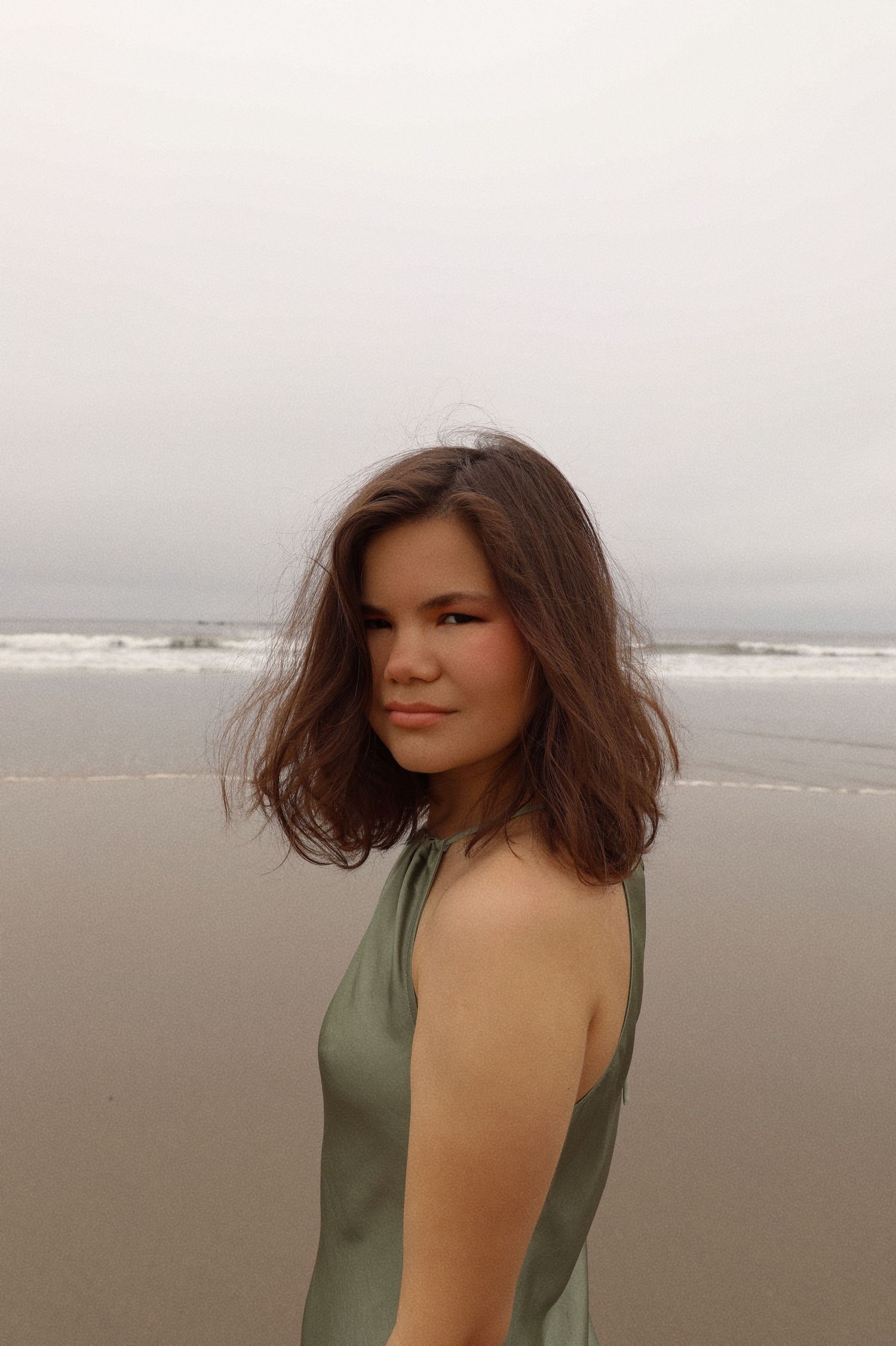 Person with wavy dark hair wearing a sage green sleeveless top poses on a cloudy beach with ocean waves in background.