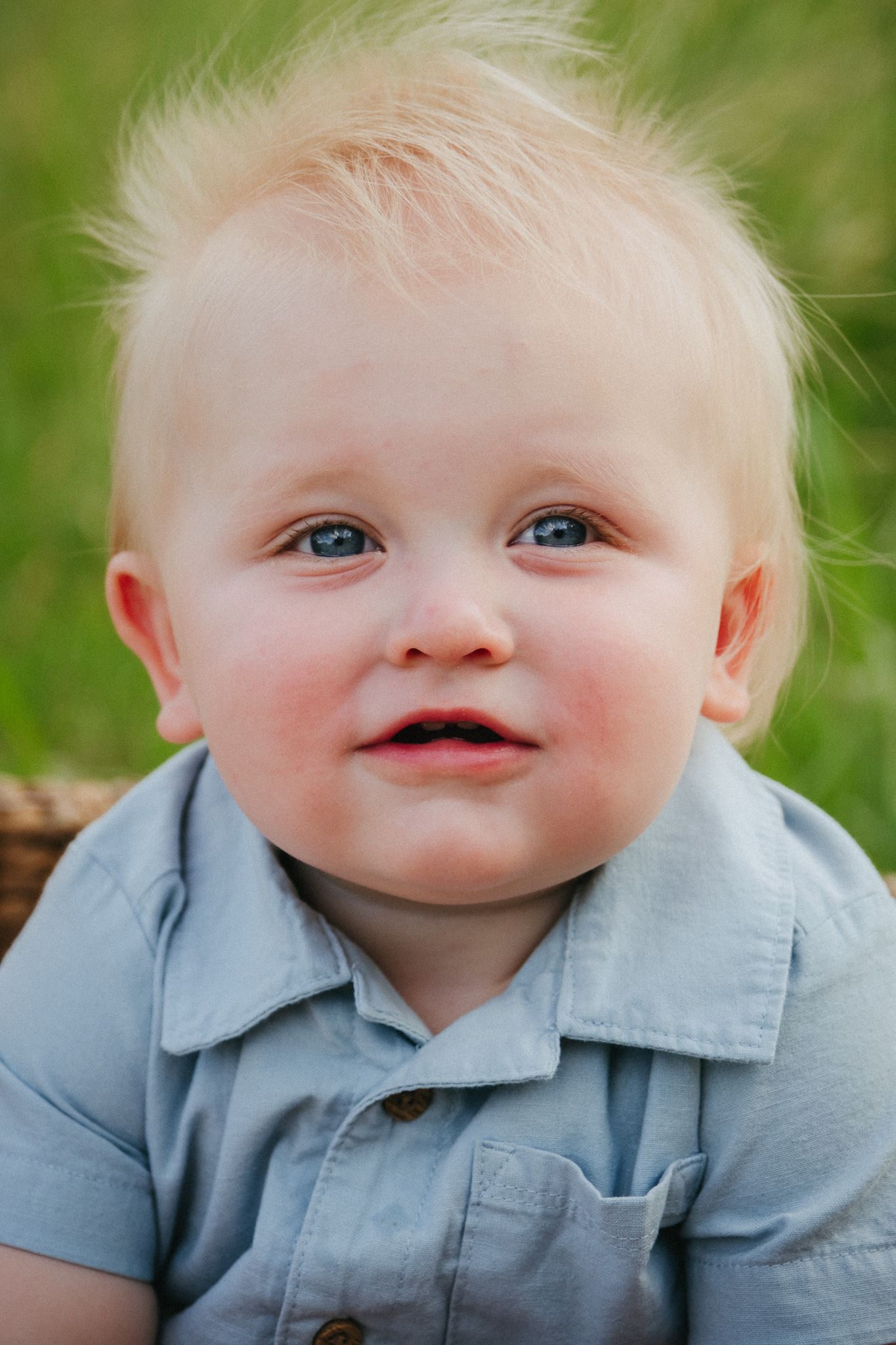 Sweet baby with light blonde hair and bright blue eyes wearing a light blue collared shirt smiles outdoors.