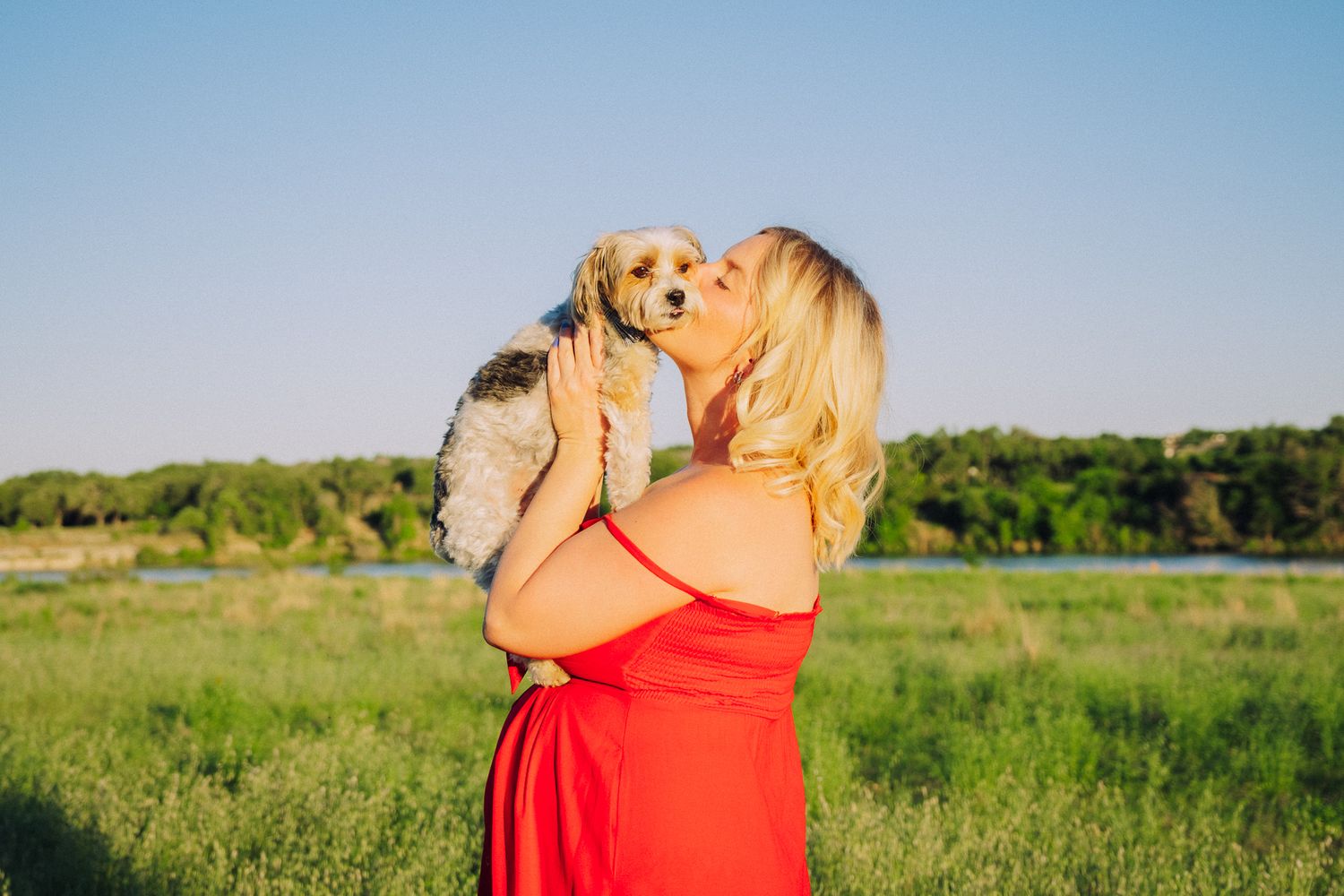 Person in a red dress holds and kisses a small golden puppy in a sunny green field.