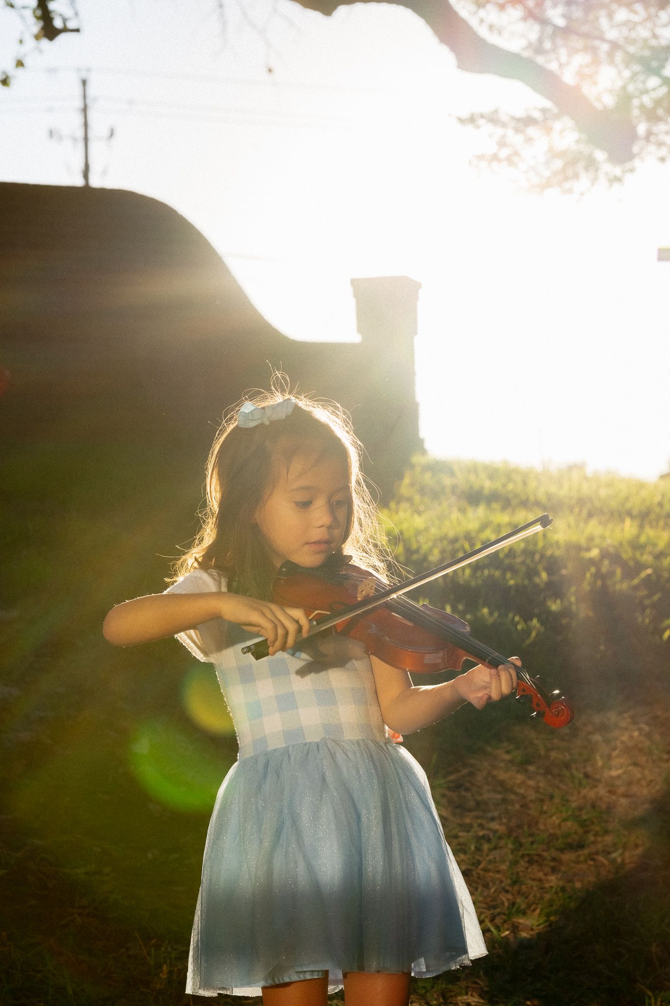 A young violinist in a blue dress practices outdoors during golden hour with sunlight creating a dreamy backlit effect.