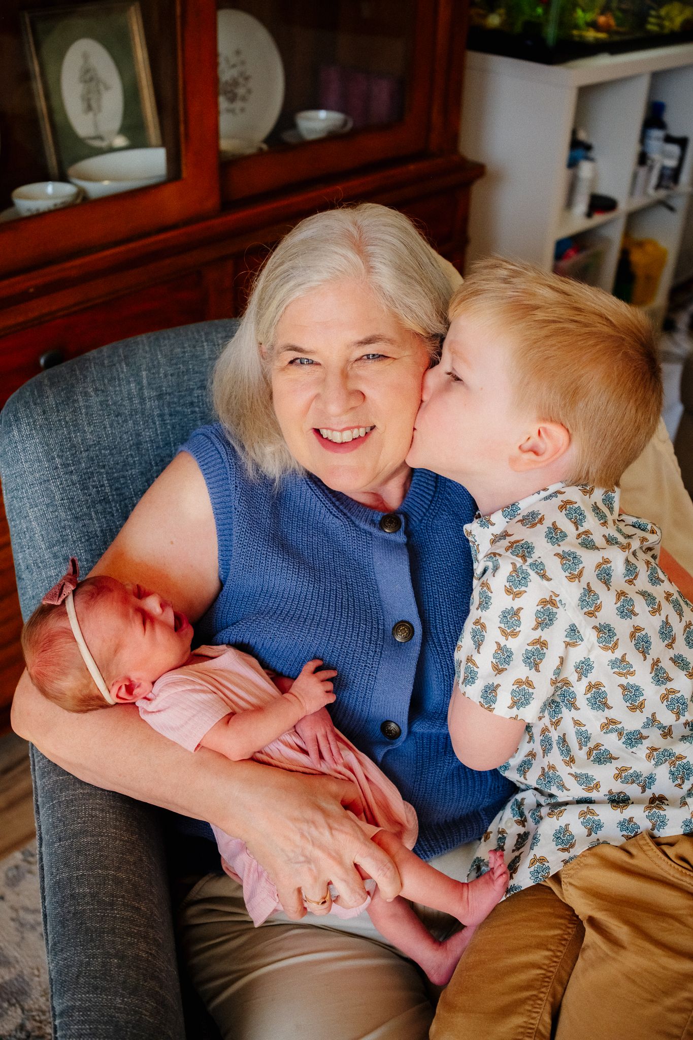 Grandmother smiling in armchair holding newborn baby girl while toddler grandson gives her a sweet kiss on the cheek.