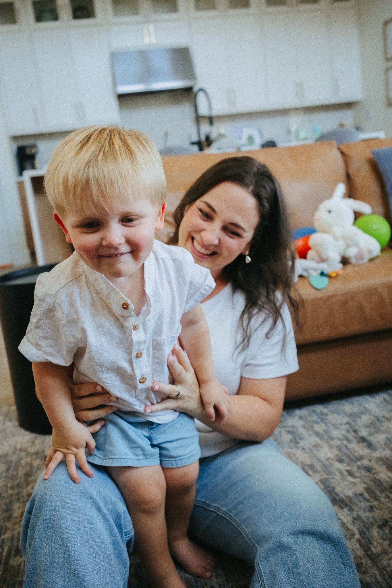 A smiling mother holds her happy blonde toddler boy on her lap in a cozy living room with toys on the sofa.