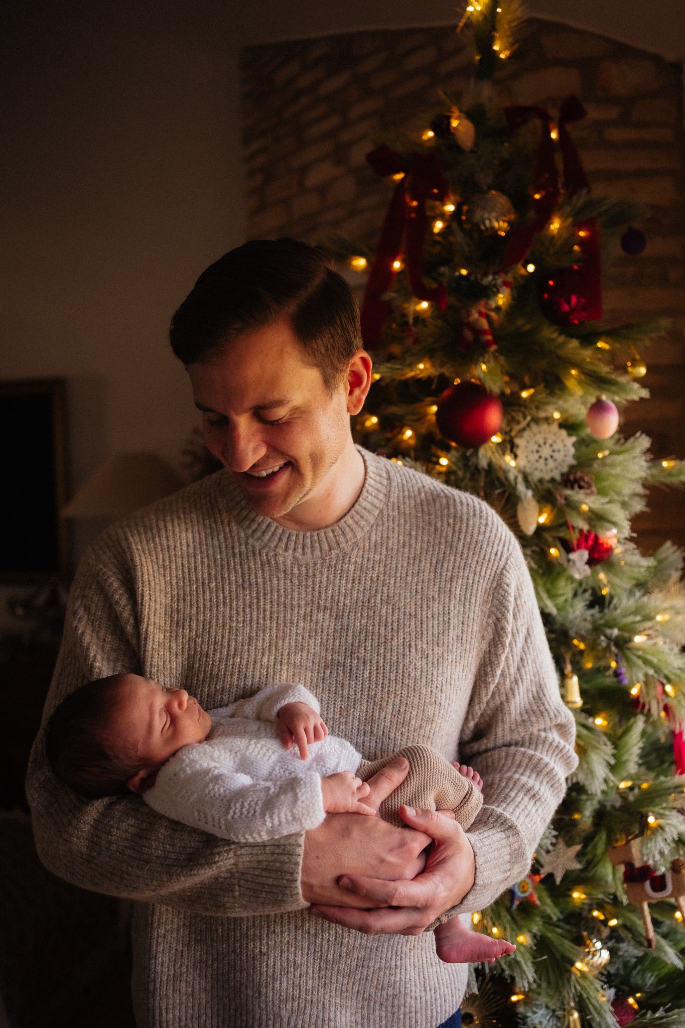 Smiling father holding newborn baby in front of decorated Christmas tree with red ornaments and warm lights.